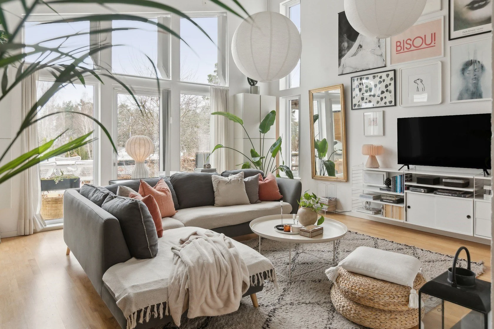 Bright living room with large windows, a gray and white sectional sofa with pink and gray pillows, a round coffee table with decor, large green plants, wall art, a TV, and a mirror.