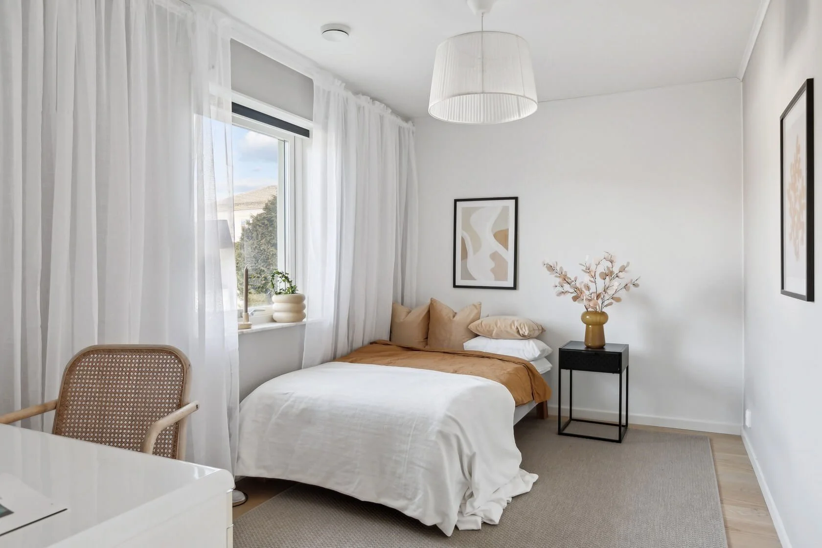 A bedroom with white walls, a bed with white and beige bedding, a black bedside table with a vase of pink dried flowers, abstract wall art, a window with white curtains, and a rattan chair.