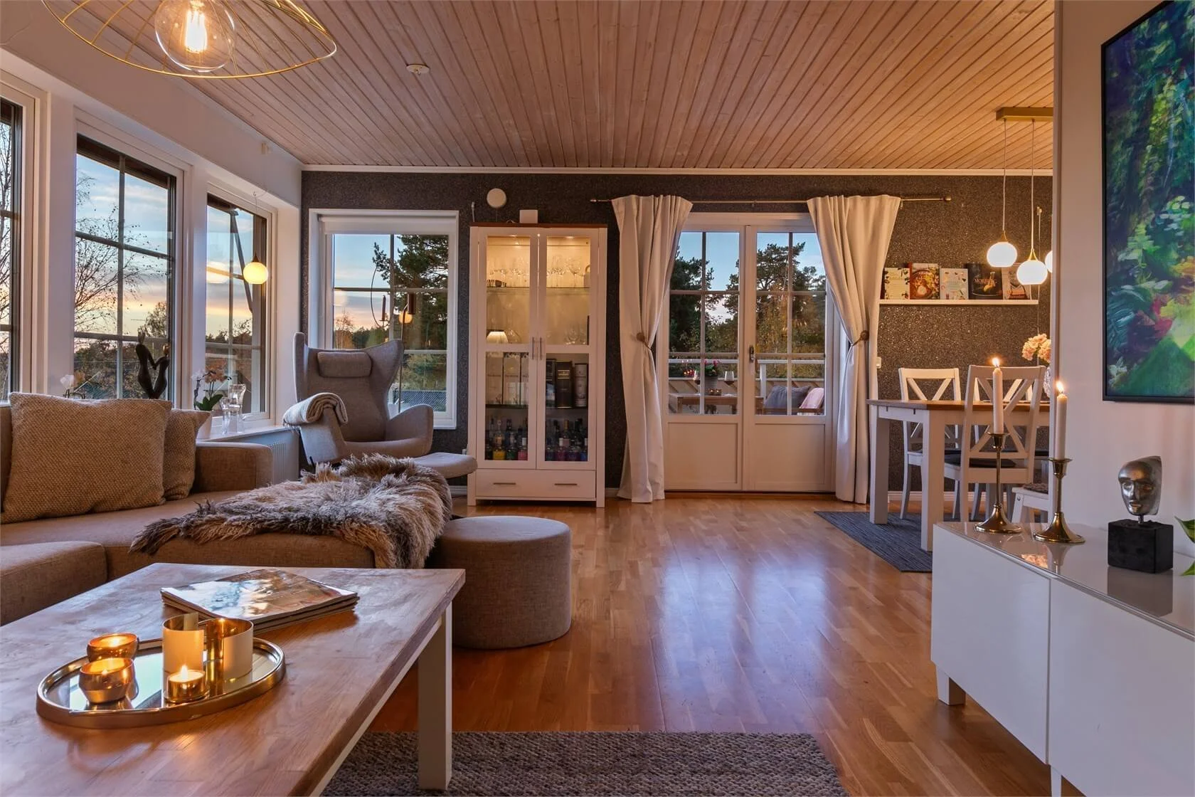 Living room with large windows showing sunset, beige sofa with pillows, furry throw, modern armchair, wooden coffee table with candles, dining area with table and chairs, wall shelf with books, and glass cabinet, hardwood floors, and warm lighting.