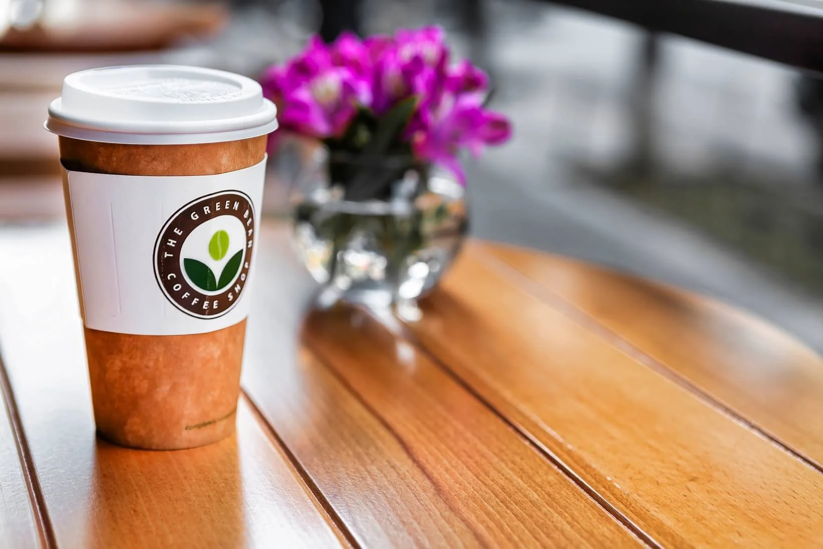 A paper coffee cup with a white lid and a logo that reads 'The Green Bean Coffee' sitting on a wooden table. A small glass vase with pink flowers is blurred in the background.