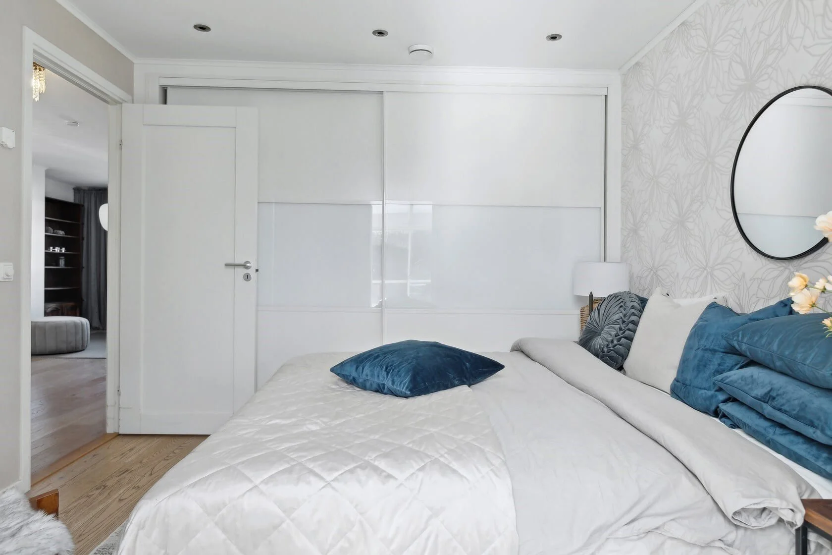 A cozy bedroom with a bed covered in white bedding and multiple blue and gray pillows, a round mirror on the wall, and a white wall with a subtle floral pattern in the background