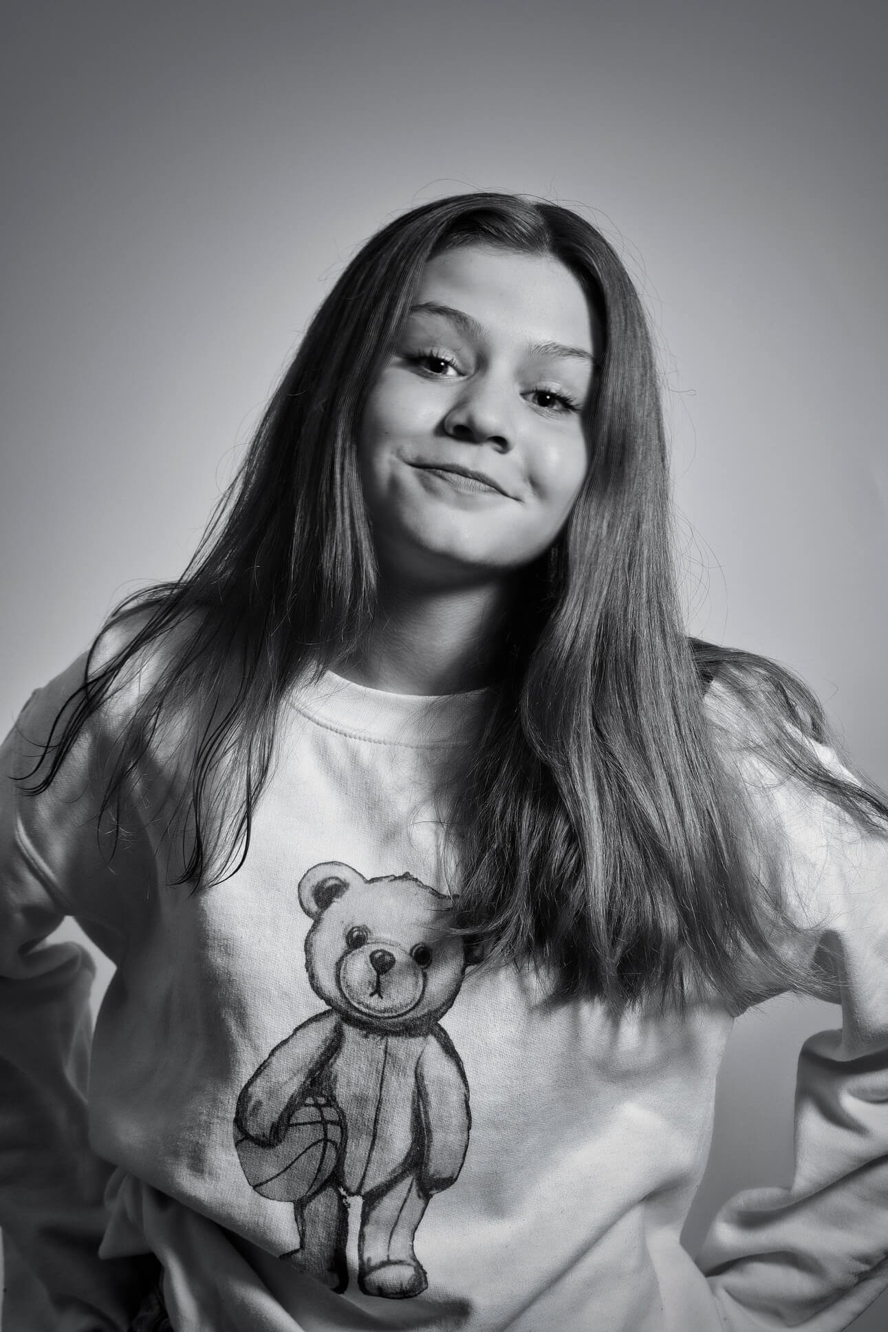 Black and white portrait of a young woman with long hair, smiling, wearing a sweatshirt with a teddy bear graphic.