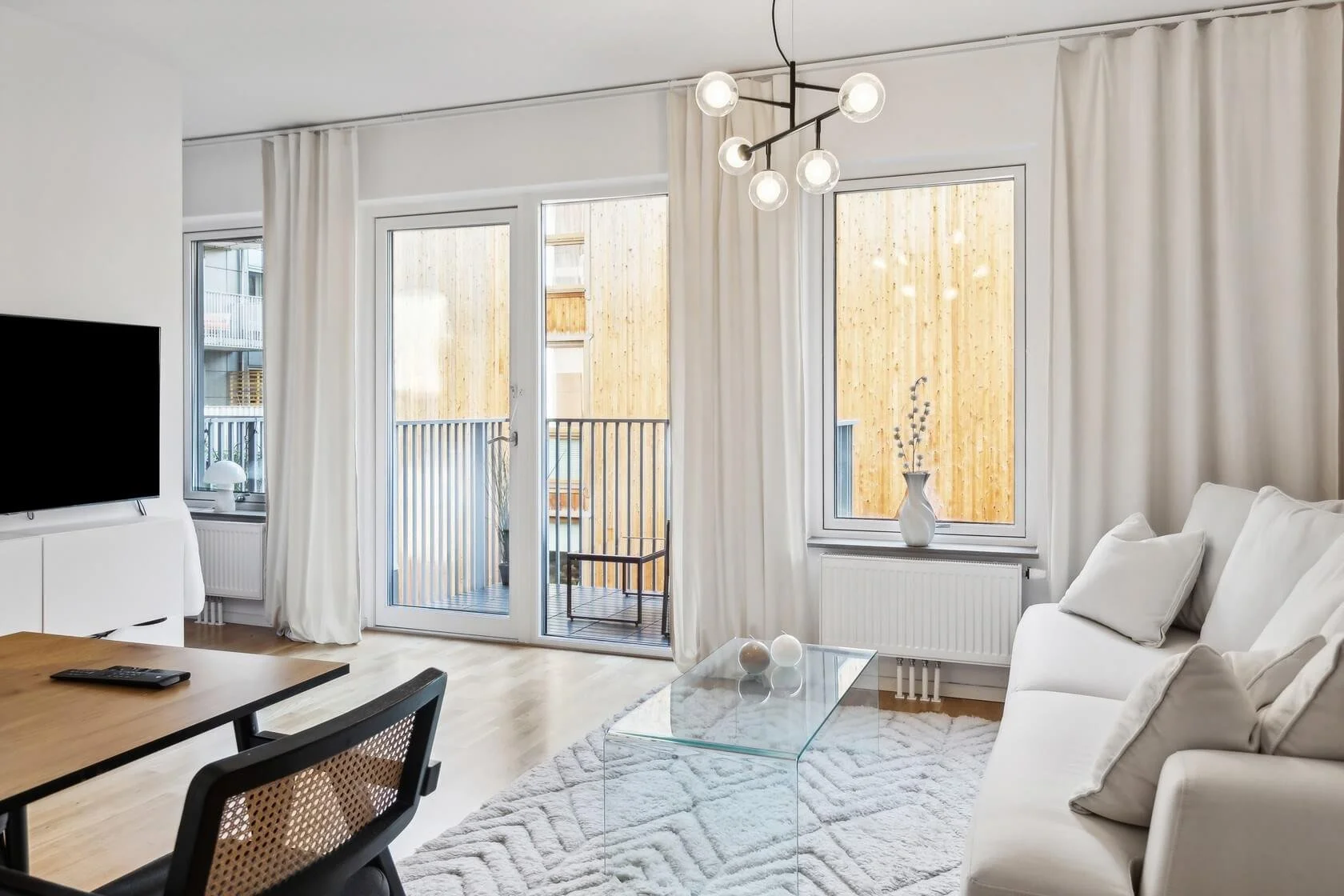 Living room with white sofa, glass coffee table, flat-screen TV, and large windows with white curtains leading to a balcony with wooden railing.