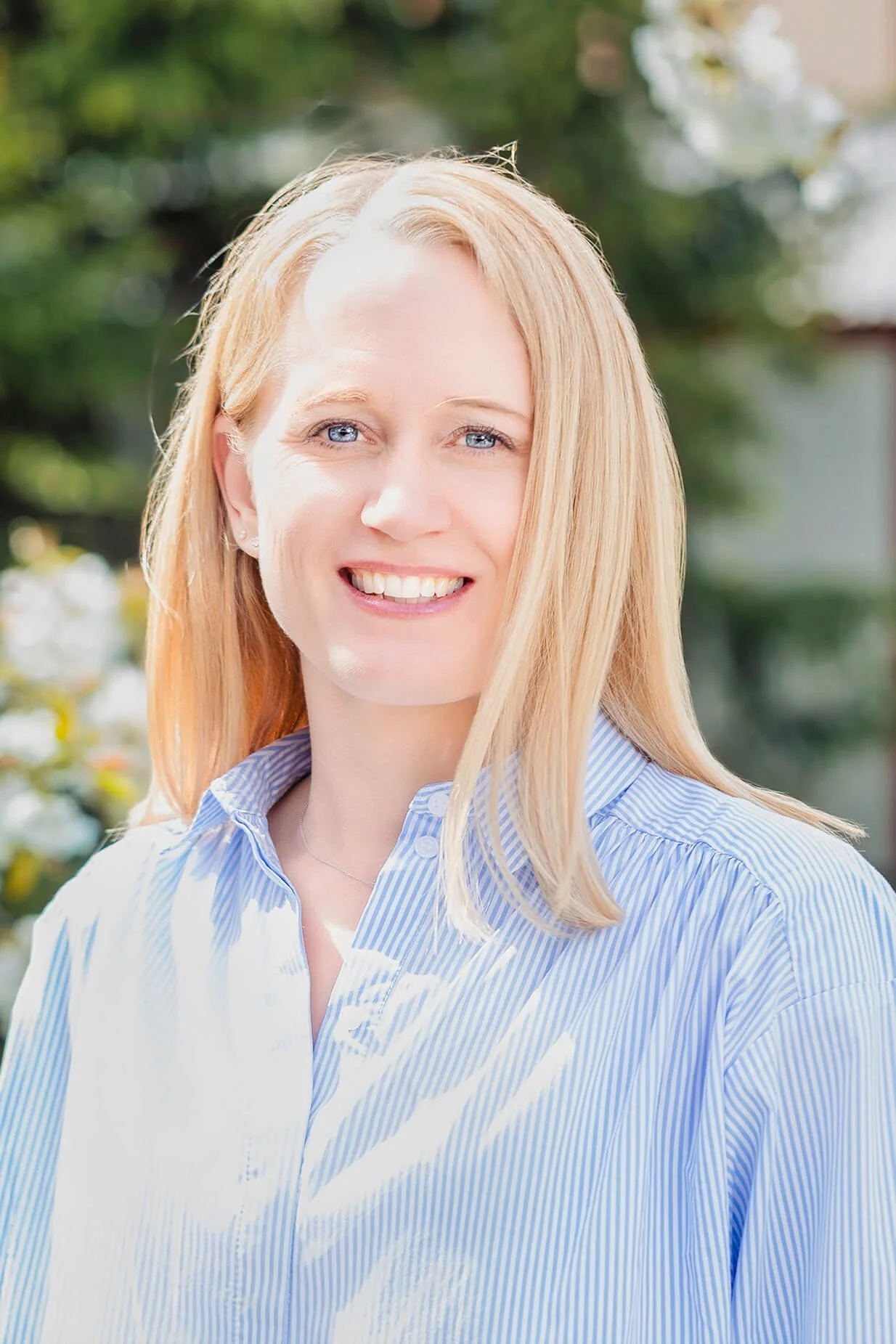 A woman with blonde hair and blue eyes smiling outdoors, wearing a blue striped shirt, against a background of greenery.