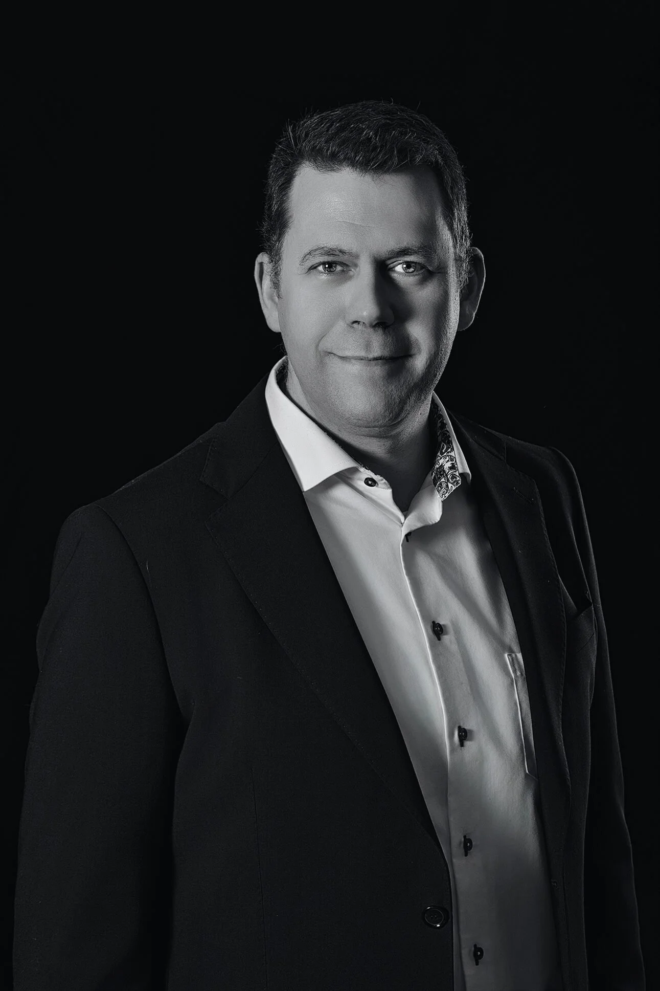 Black-and-white professional portrait of a man in a suit, smiling slightly, against a dark background.