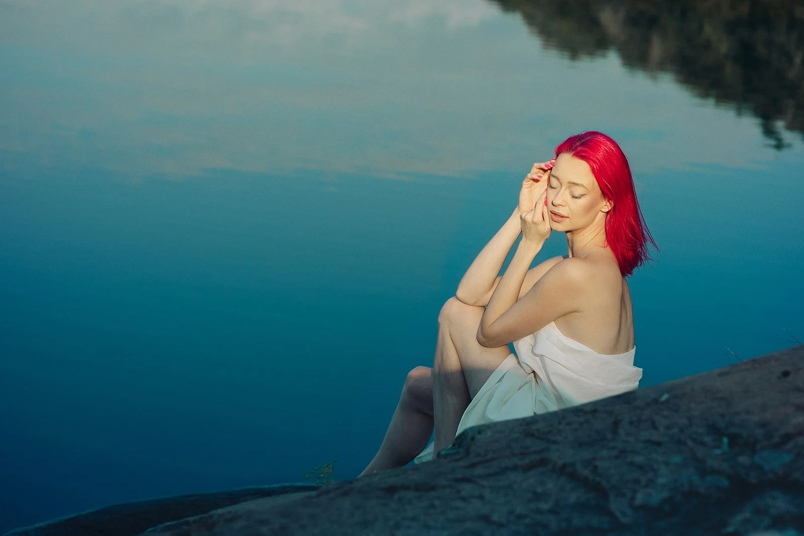 A woman with red hair sitting on a rock by a body of water, eyes closed, holding her face with one hand, wearing a white cloth over her shoulders.