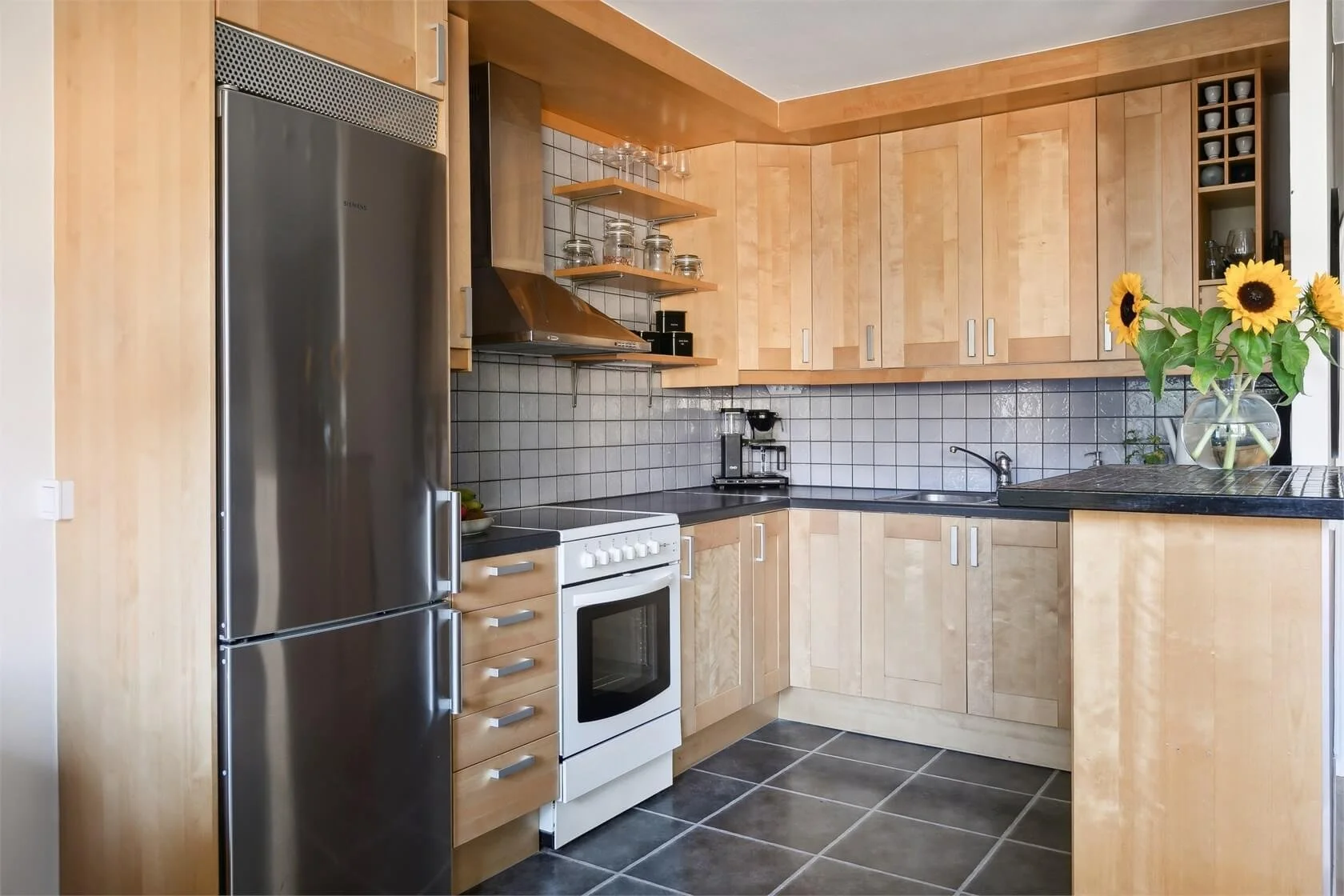 Kitchen with wooden cabinets, black countertops, stainless steel refrigerator, white stove, and a black tile backsplash. There is a sunflower bouquet in a glass vase on the counter.