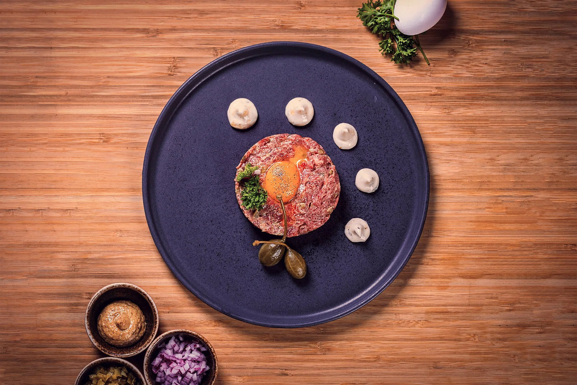 Raw ground beef patty topped with a raw egg yolk, garnished with parsley and caper berries on a blue plate, with dollops of mayonnaise and chopped vegetables surrounding it, on a wooden surface.
