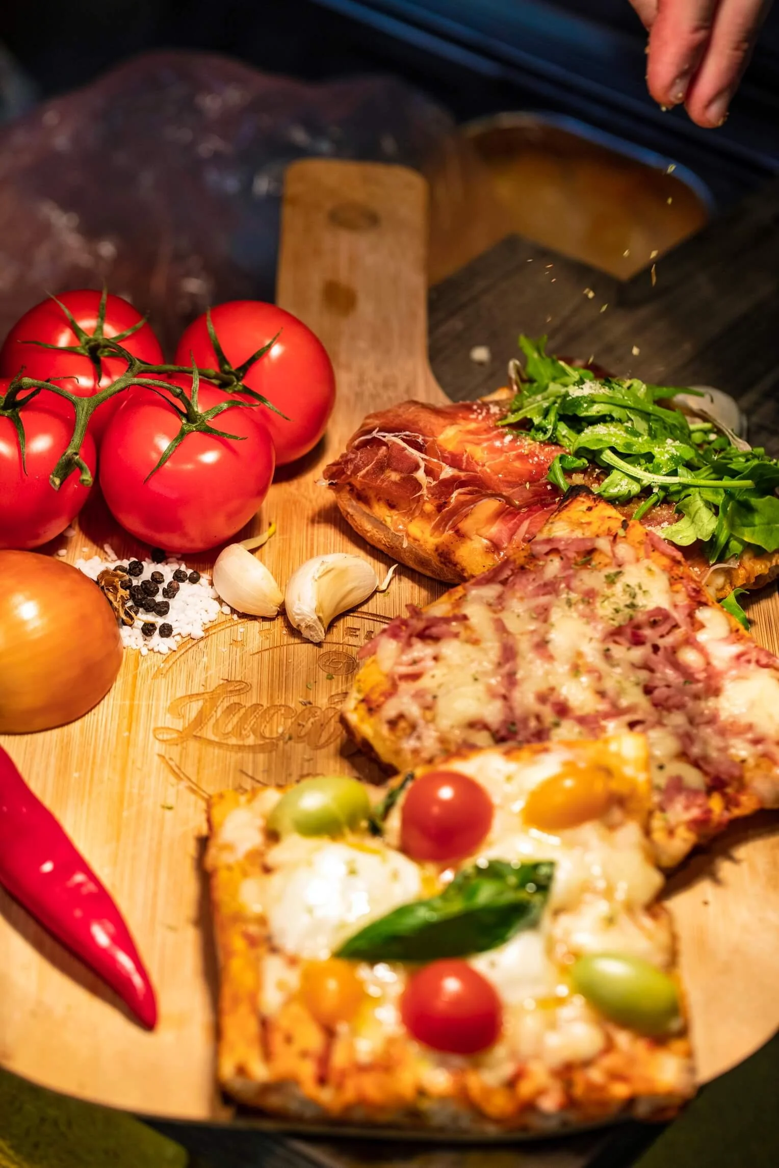 A wooden board holds a sliced pizza with cherry tomatoes, basil, and cheese, with anchovies and herbs. Nearby, there are fresh tomatoes on the vine, garlic cloves, a yellow onion, a red chili pepper, black peppercorns, and salt.