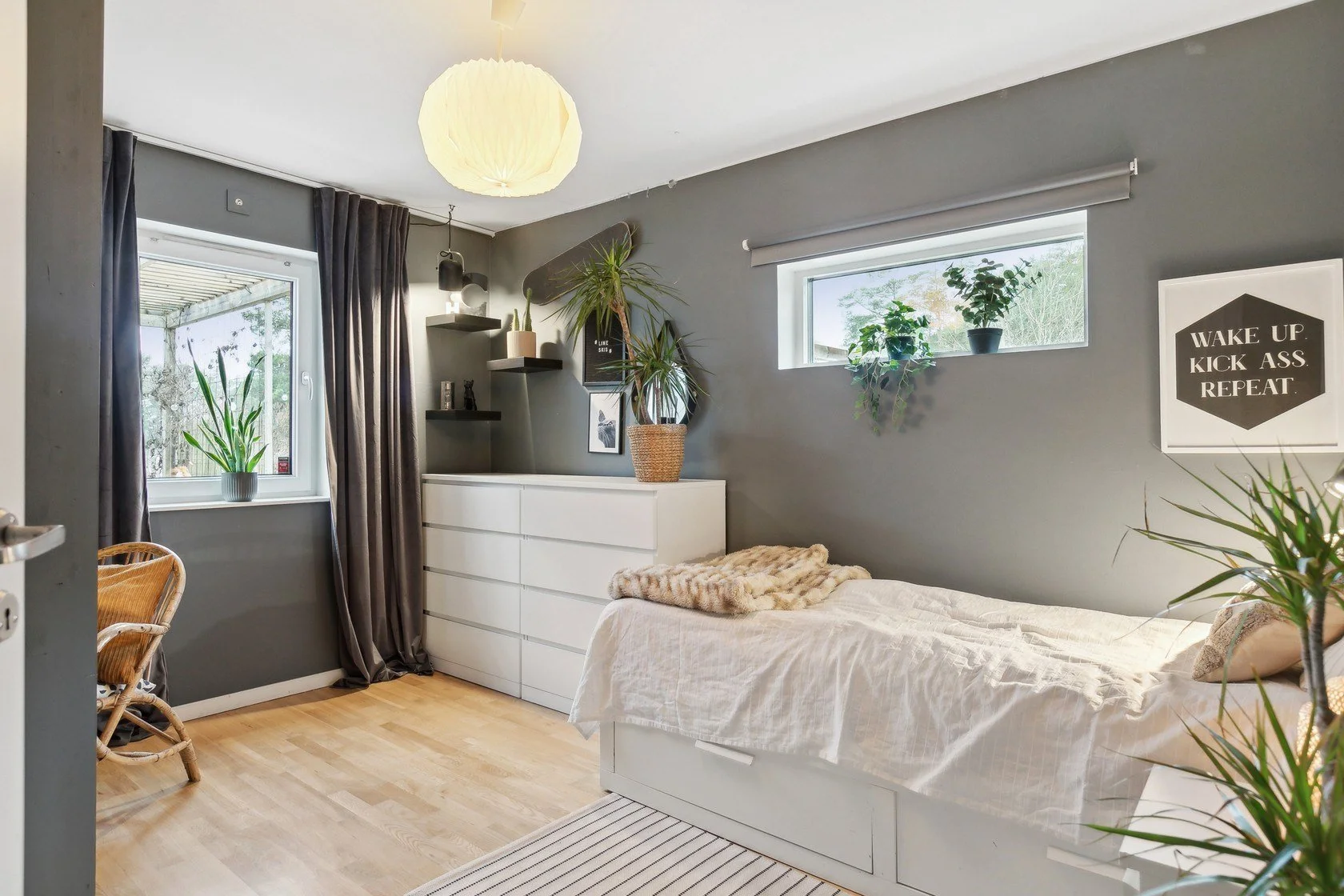 A bedroom with gray walls, a white bed with beige bedding, and green plants on the windowsill and on a dresser. There are black curtains, a wicker chair, and wall art including a black and white portrait and a sign that reads 'WAKE UP. KICK ASS. REPE