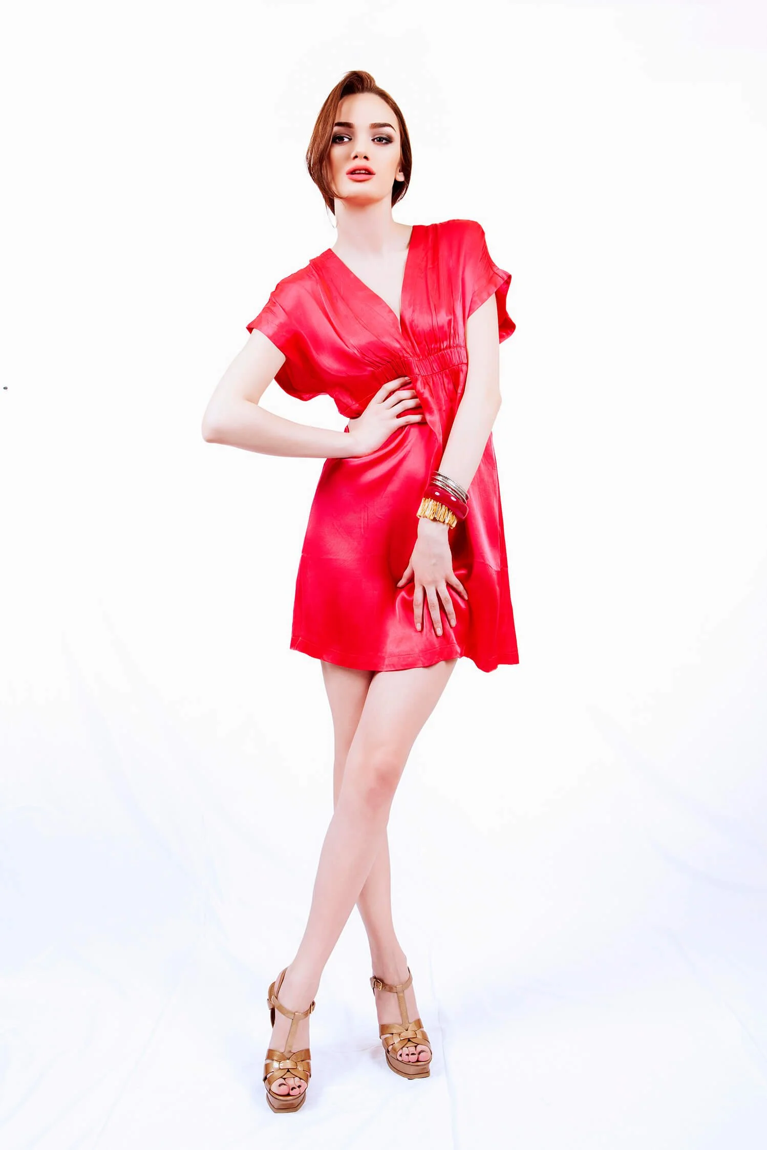 A woman with shoulder-length hair in a red satin dress and high-heeled sandals standing against a white background.