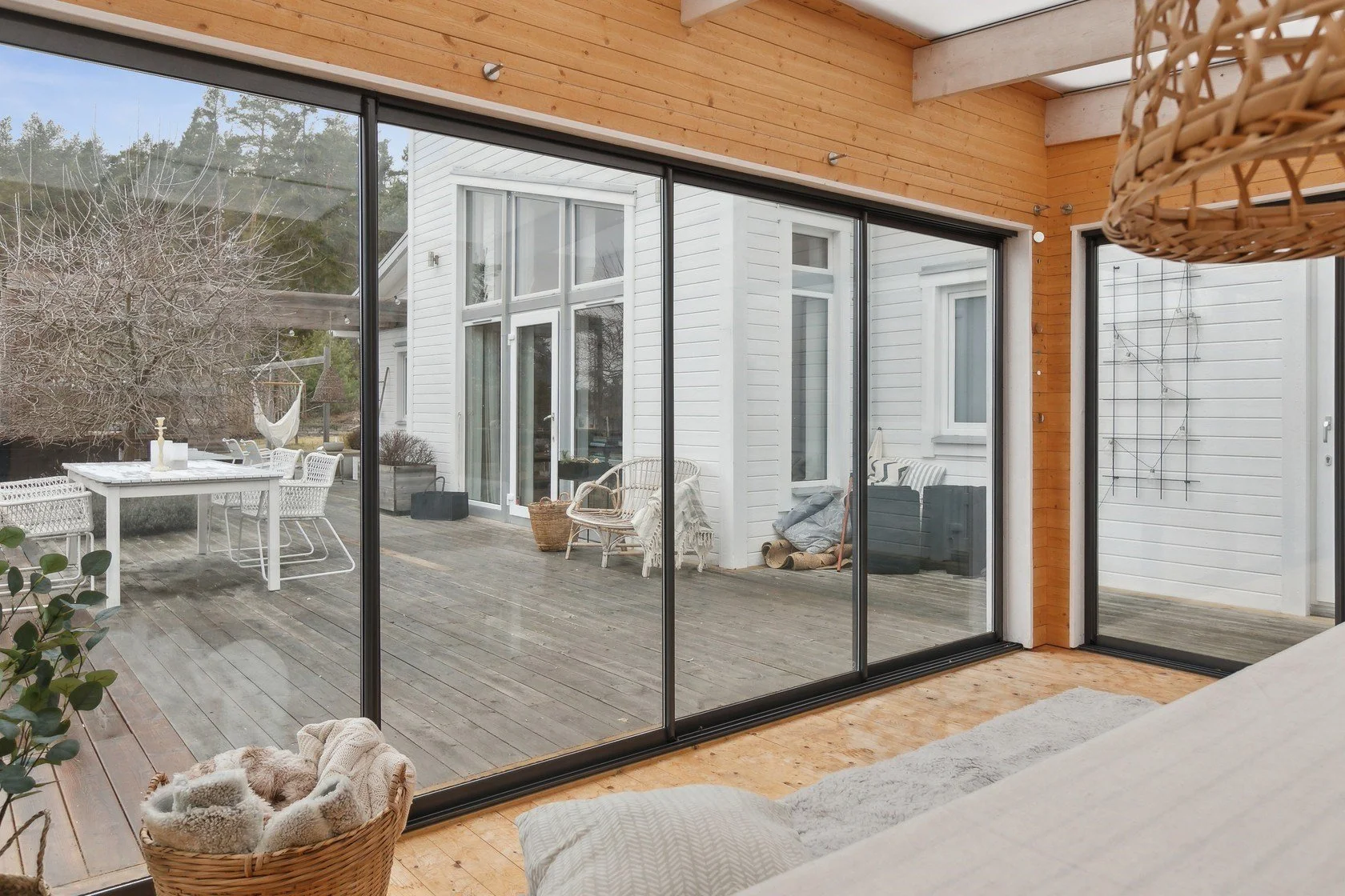 View of a wooden deck outside a house, seen through large sliding glass doors from a cozy indoor space with a wicker basket filled with blankets and a bed with neutral-colored bedding.