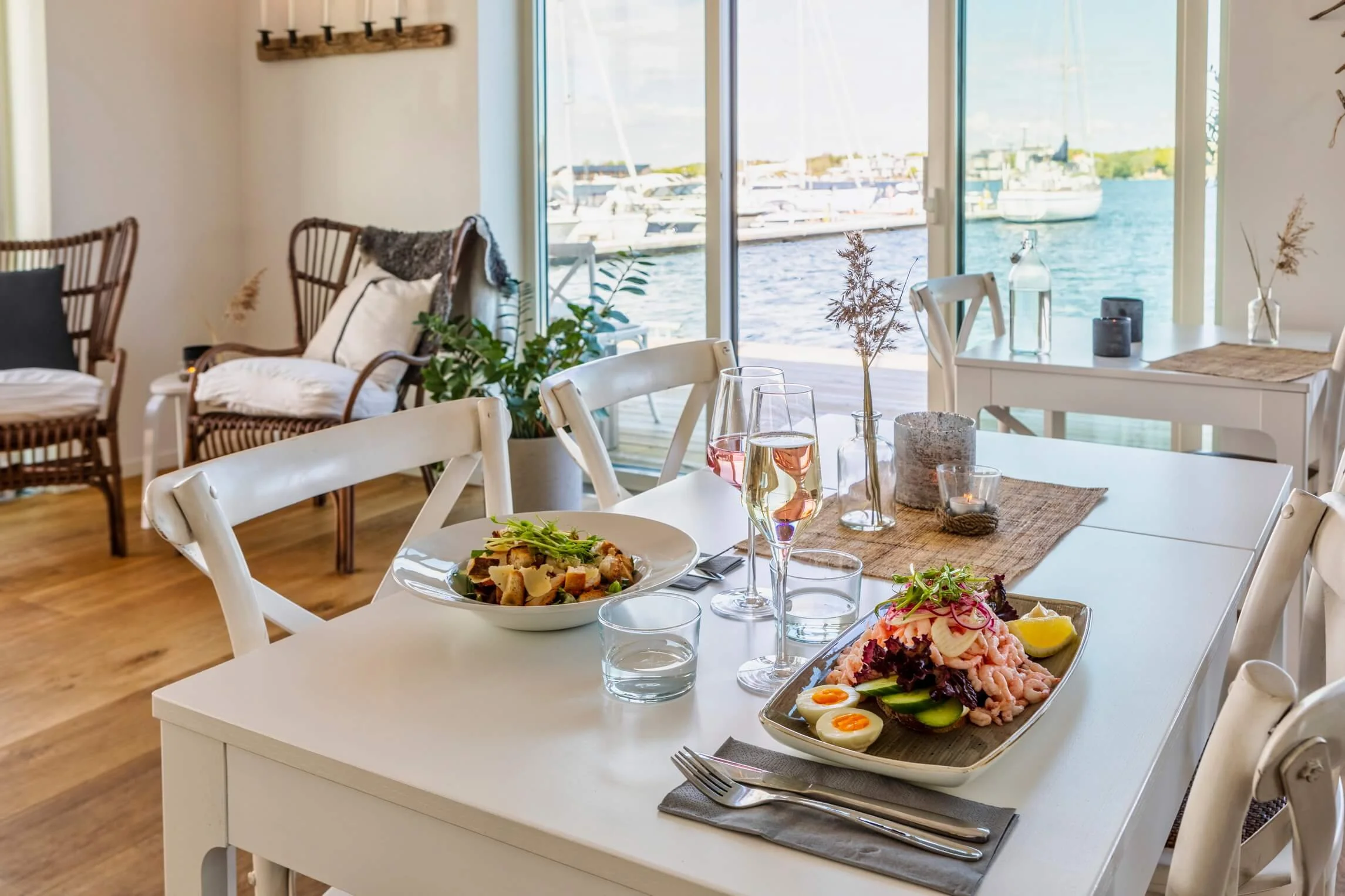 Dining table set with salads, glasses of wine and water, in a bright room with large windows and a view of boats on a waterway.