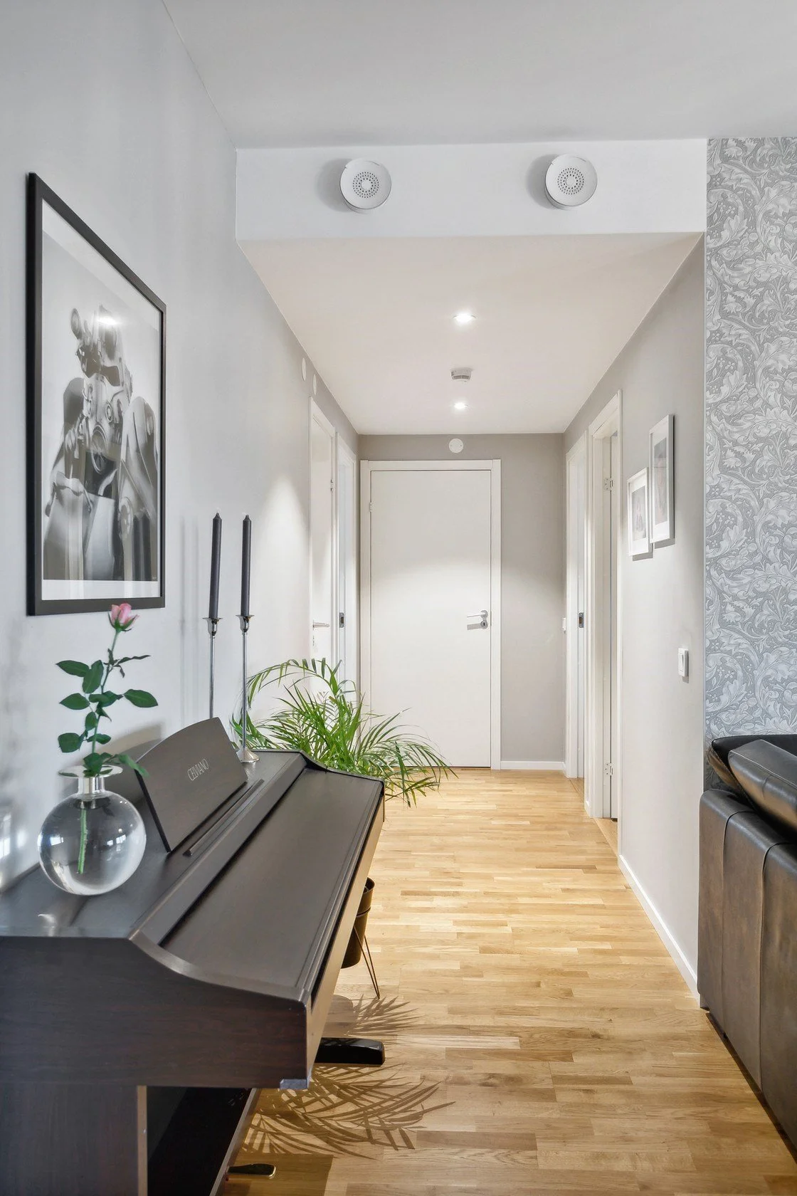 A hallway with wood flooring, white walls, decorated with framed photographs and artworks. A black digital piano with a glass vase containing a pink rose and two black candles on top. A green potted plant. Doors leading to other rooms, and a textured