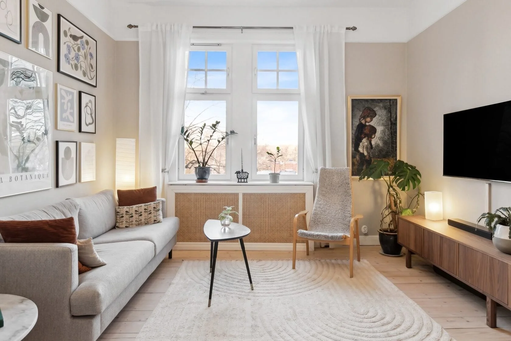 Living room with a light gray sofa, decorative pillows, a small black coffee table, a textured armchair, a wooden TV stand with a mounted flat-screen TV, houseplants, art on the walls, a large window with white curtains, and a beige circular rug.