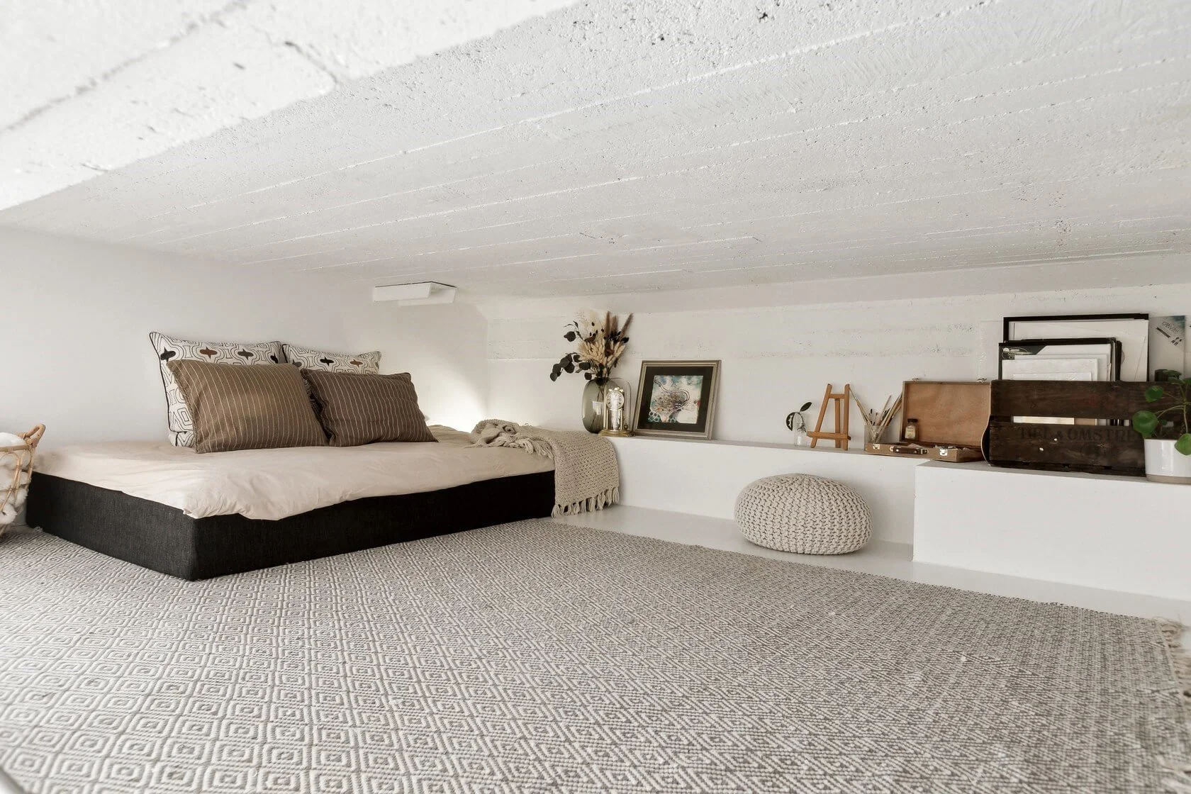 Minimalist bedroom with low bed, neutral-toned pillows, a textured cream blanket, decorative vase with dried flowers, framed artwork, small wooden easel, storage boxes, potted plant, knit pouf, and patterned rug on white walls and ceiling.