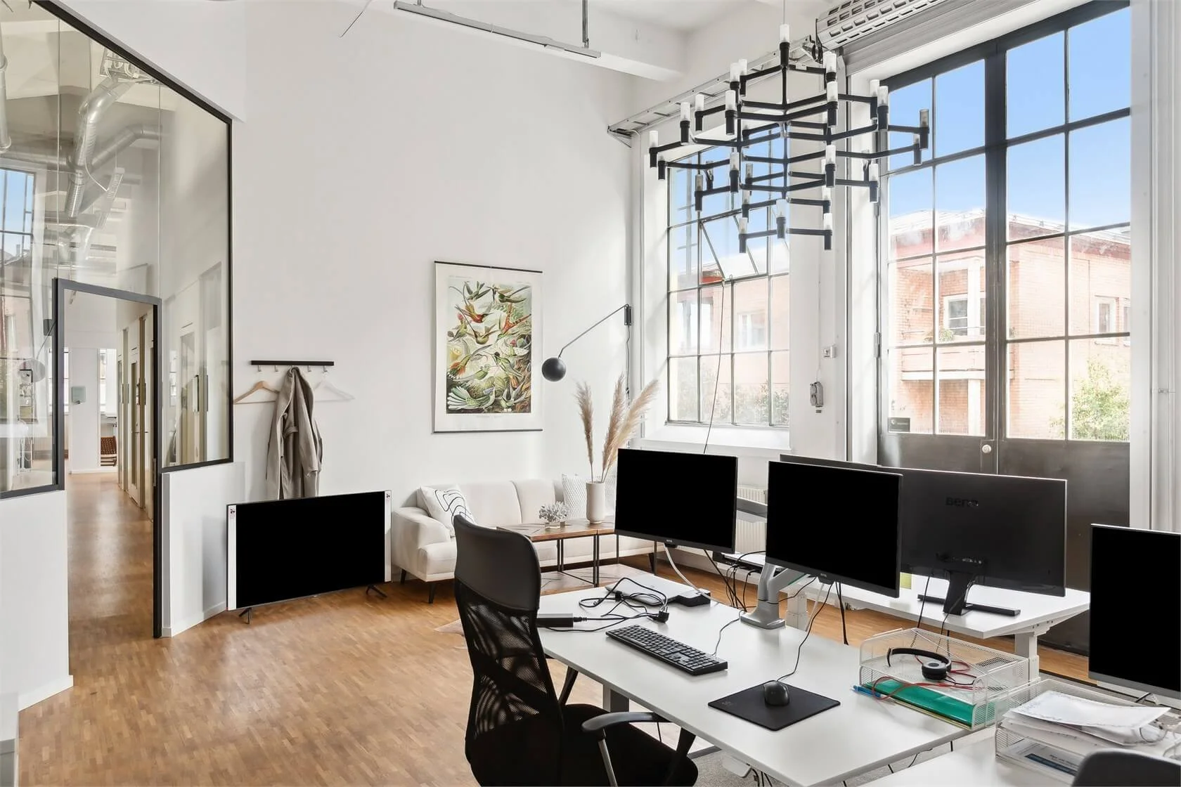 Modern office space with white walls, large windows, multiple computer monitors on a white desk, black office chair, a white sofa, a framed botanical print on the wall, and a black geometric chandelier overhead.