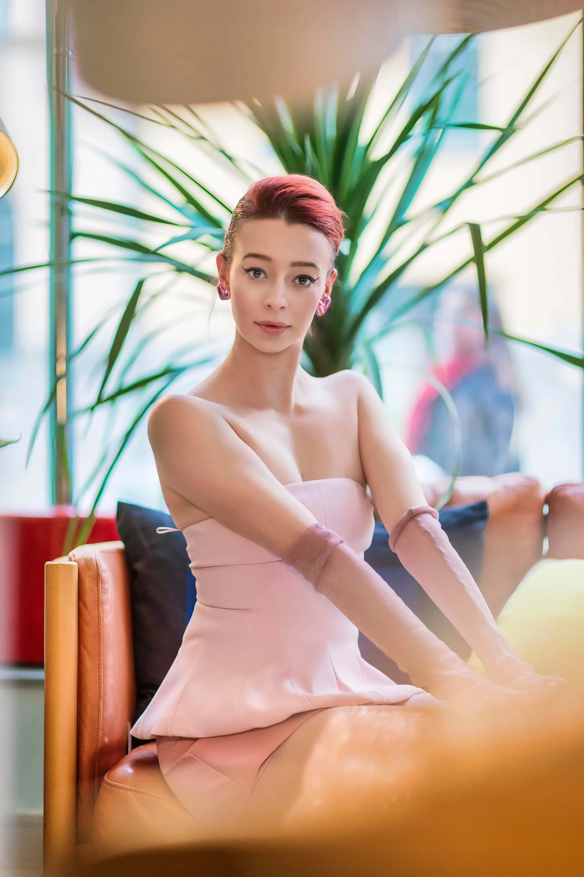 A woman with red hair styled in an updo, wearing a strapless pink dress and long pink gloves, sitting on a chair in a cafe or restaurant with large green plants and colorful cushions in the background.
