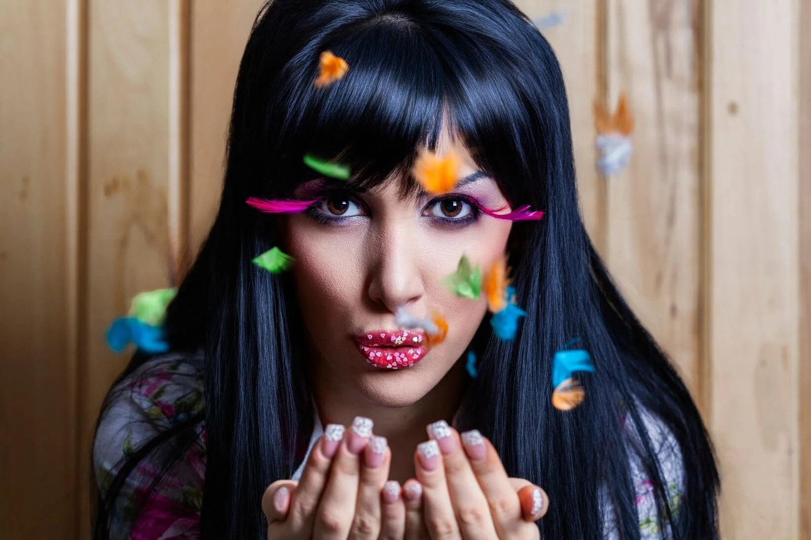 Woman with long black hair and colorful makeup blowing small, colorful paper confetti while standing in front of a wooden wall.