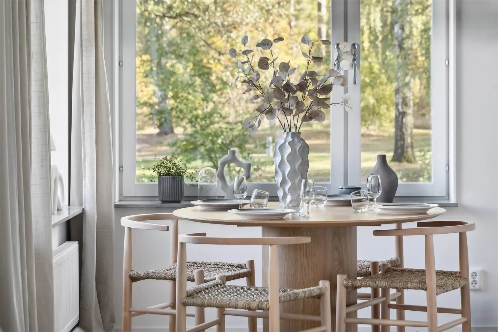 Dining area with a round wooden table set with white plates and glasses, decorated with a tall white vase filled with eucalyptus branches, surrounded by three wooden chairs with woven seats, next to a large window with white curtains overlooking a gr