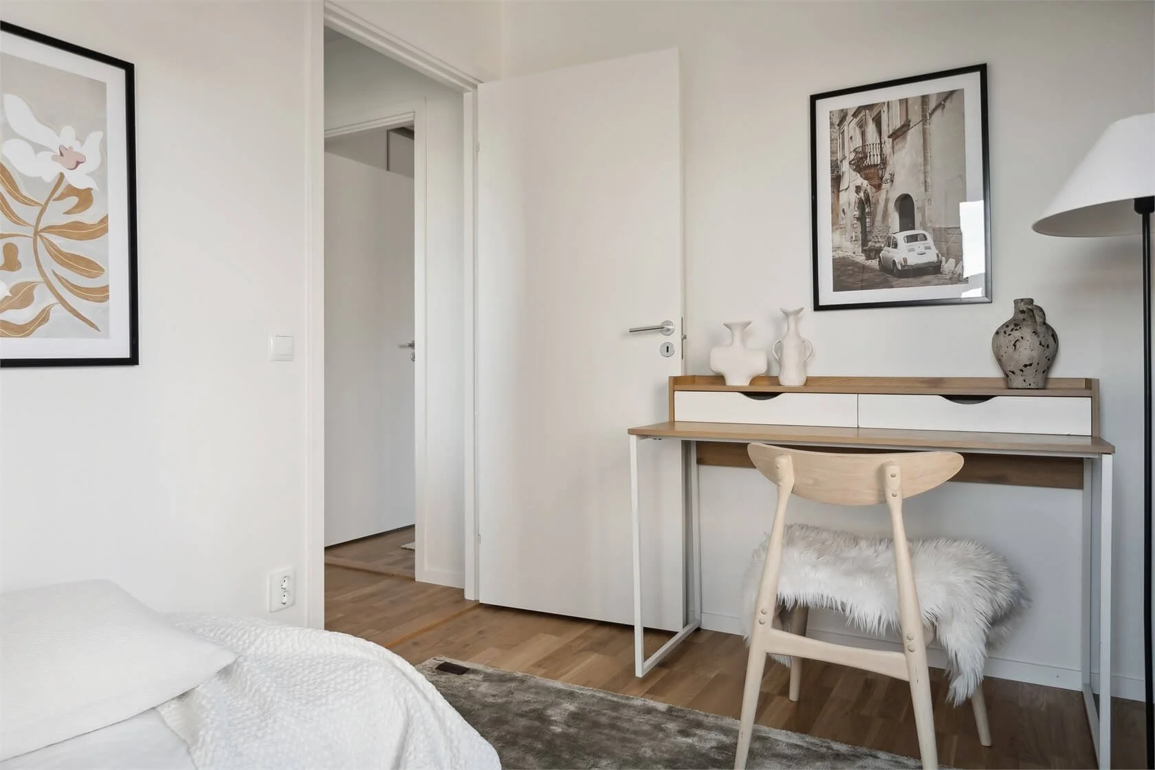 A minimalist bedroom corner with a white bed, a wooden desk, a wooden chair with a furry cushion, framed black and white artwork on the walls, a tall standing lamp, and decorative vases on the desk.