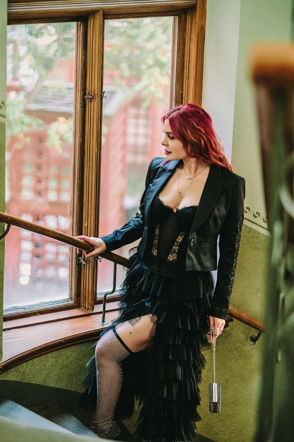 Woman with red hair in black fashion standing on a staircase by a large window, holding a handrail, wearing fishnet stockings, a black lace corset, and a black jacket with ruffled skirt, holding a small clutch purse.