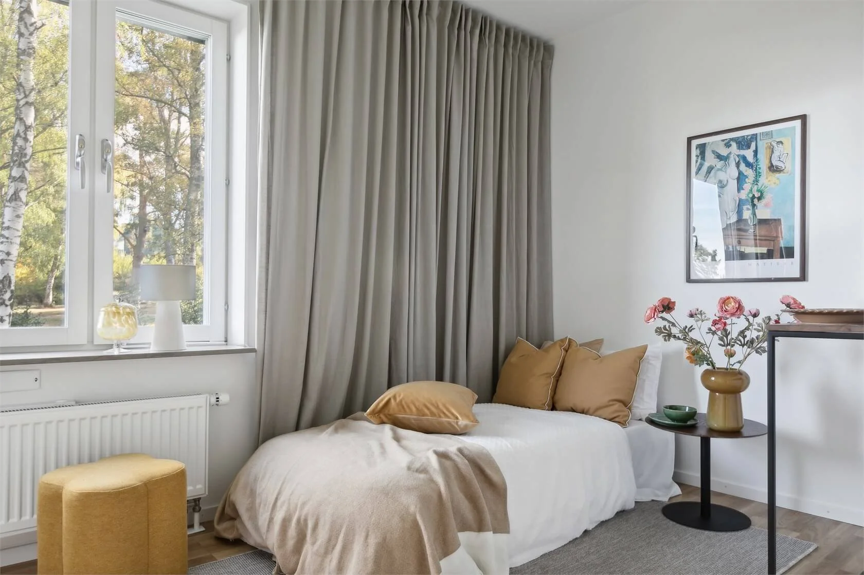 A cozy bedroom corner with a white bed, beige pillows, and a blanket, beside a window with cream curtains. A black side table with pink flowers in a yellow vase, a green bowl, and art on the wall.
