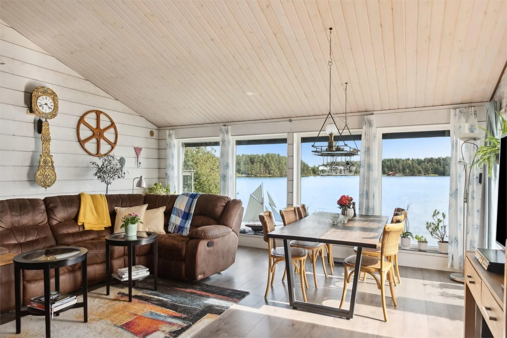Living room with large windows overlooking a lake, featuring a brown leather sofa, a wooden dining table with six chairs, potted plants, and decorative wall items, including a clock and a wheel, with sunlight streaming in.
