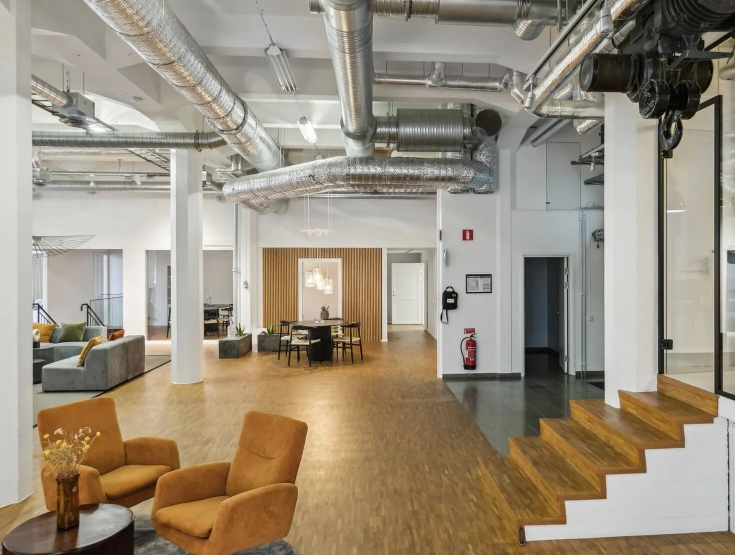 Interior of a modern office or common area with exposed ductwork, wooden flooring, and contemporary furniture including chairs, sofas, and a dining table with chairs.