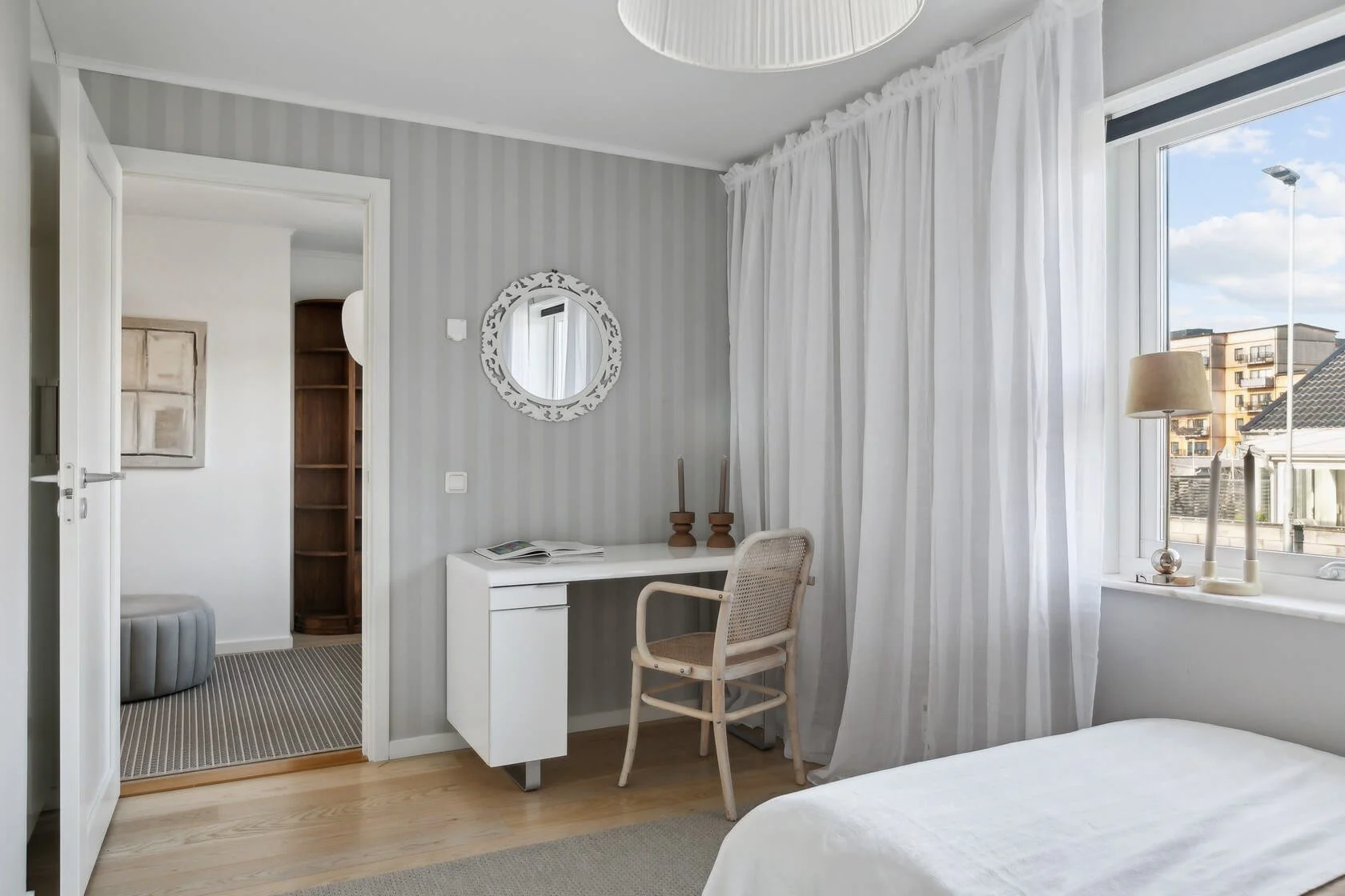 A bedroom with white curtains, a bedside lamp, a small white desk, an armless chair, and a window showing a view of neighboring buildings