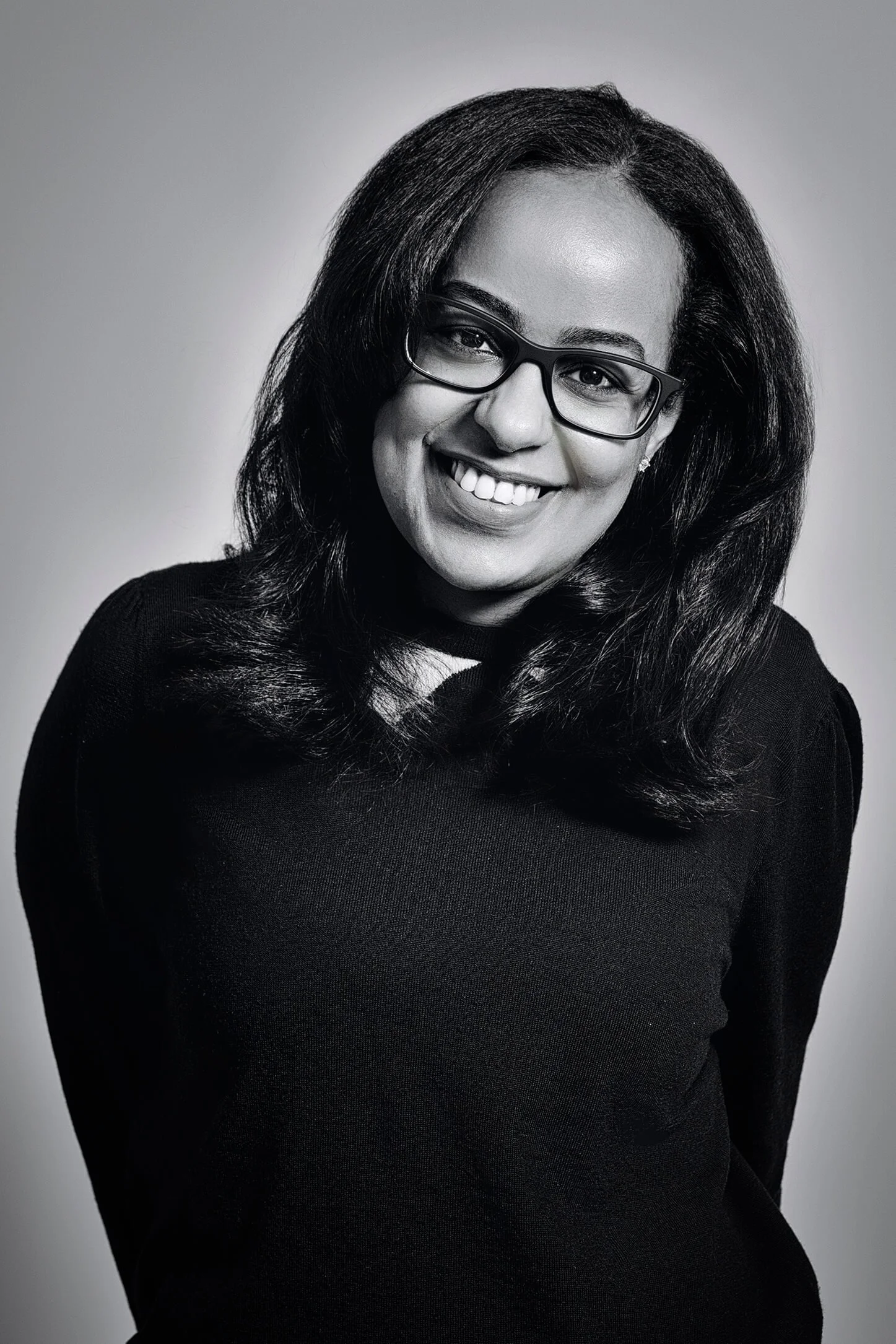 A woman with dark, shoulder-length hair, glasses, wearing a black top, smiling in a portrait photograph.