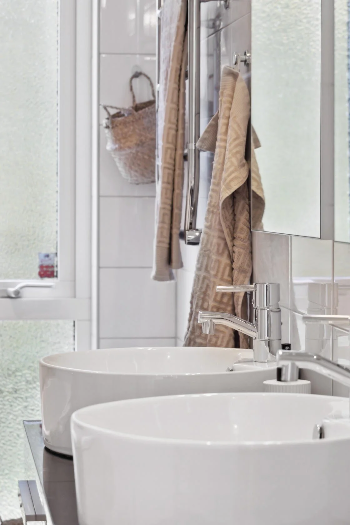 A bathroom with two white sinks, beige towels hanging on a rack, a mirror, a window with frosted glass, and a woven basket on a tiled wall.