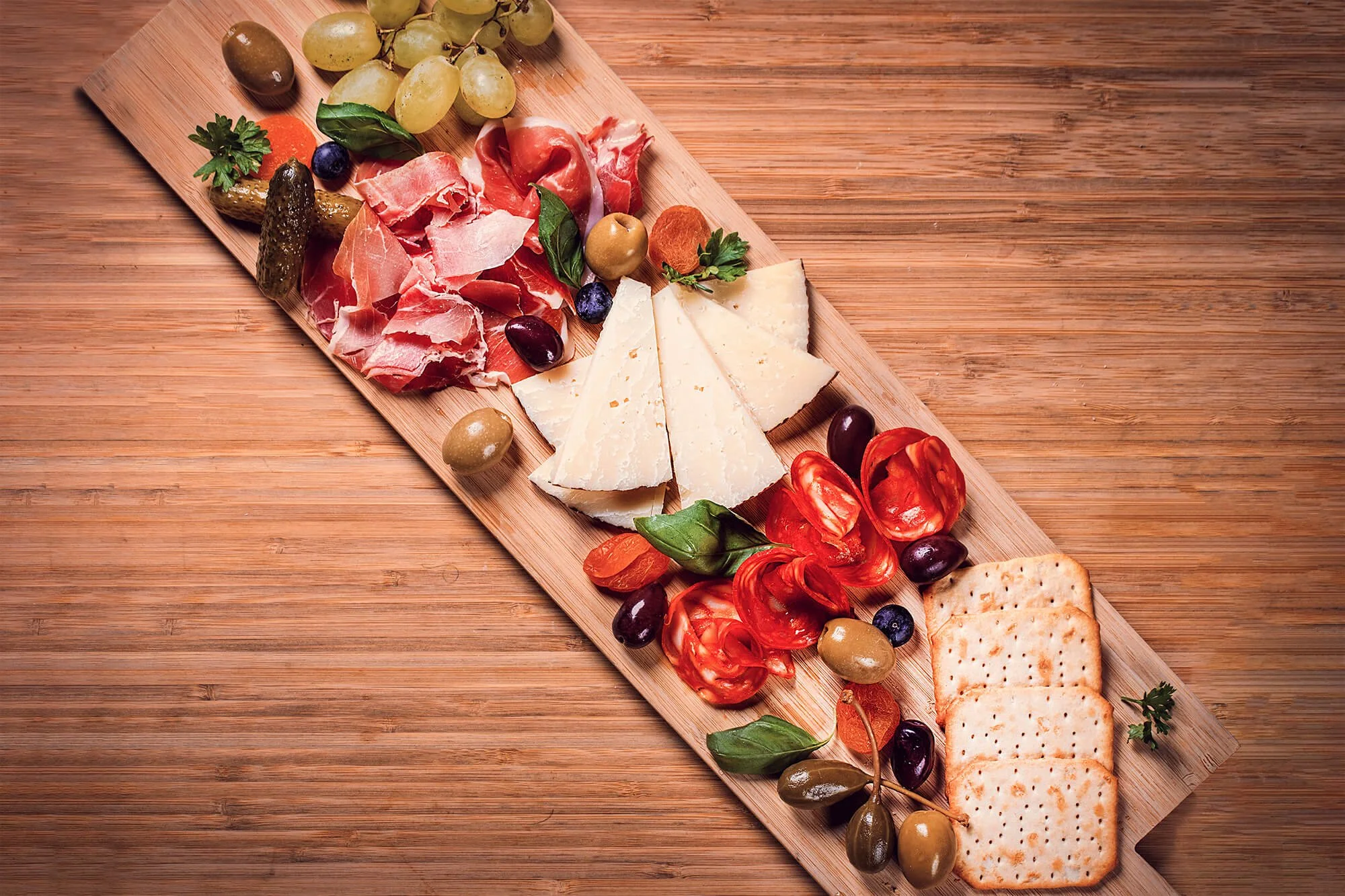 A wooden serving board with cheese, cured meats, grapes, crackers, and olives. The board is placed on a wooden surface.