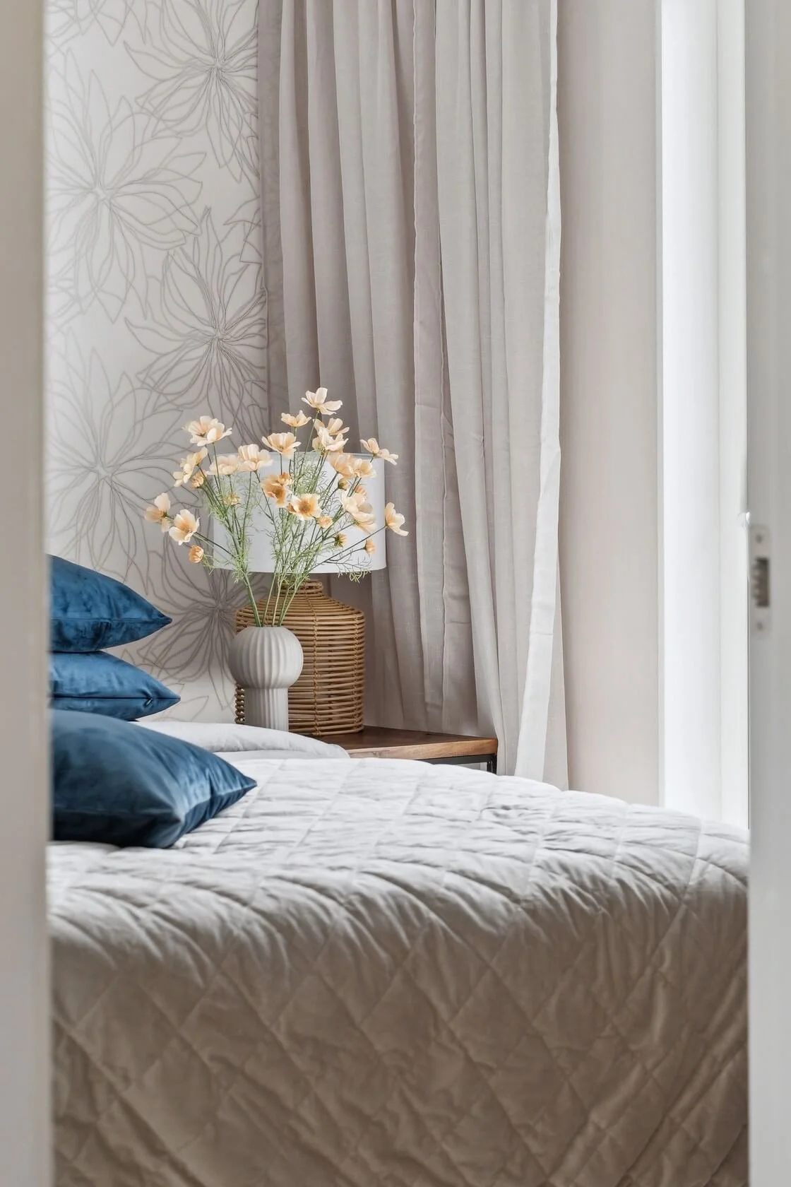 A cozy bedroom corner with a bed made with white quilted bedding and three dark blue pillows. A nightstand with a decorative vase of white and peach flowers, a wicker table lamp, and beige curtains next to a sheer curtain cover the window.