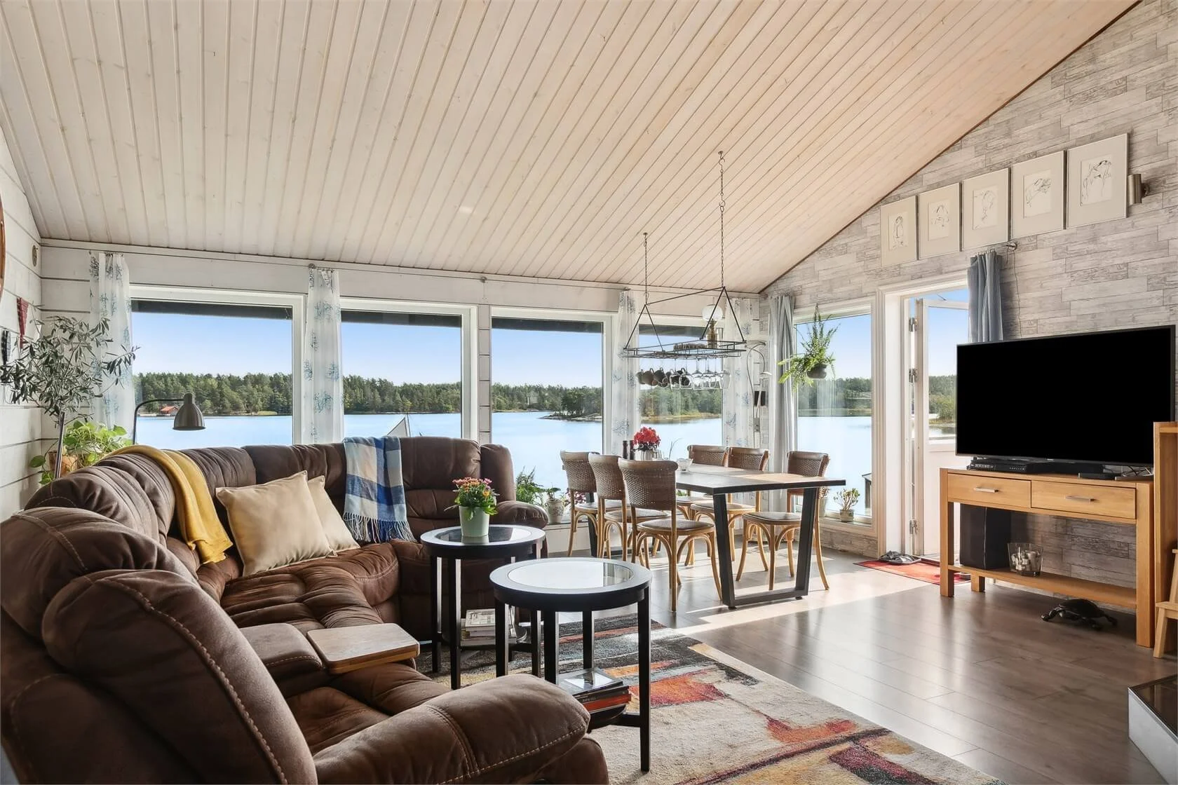 Living room with large windows overlooking a lake, featuring a brown sectional sofa, coffee tables, a dining table with chairs, a television on a wooden stand, various plants, and decorative elements.