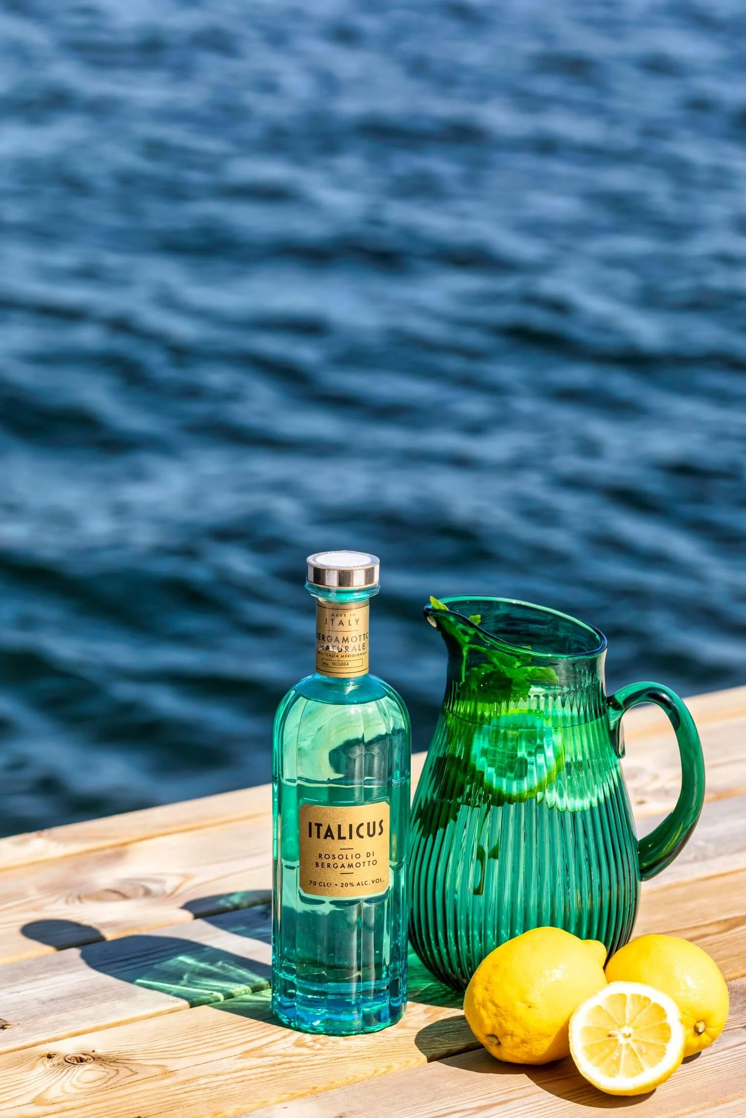 A bottle of Italicus Rosolio di Bergamotto, a green ribbed glass pitcher with a lemon slice inside, and two whole lemons, placed on a wooden surface near a body of water.