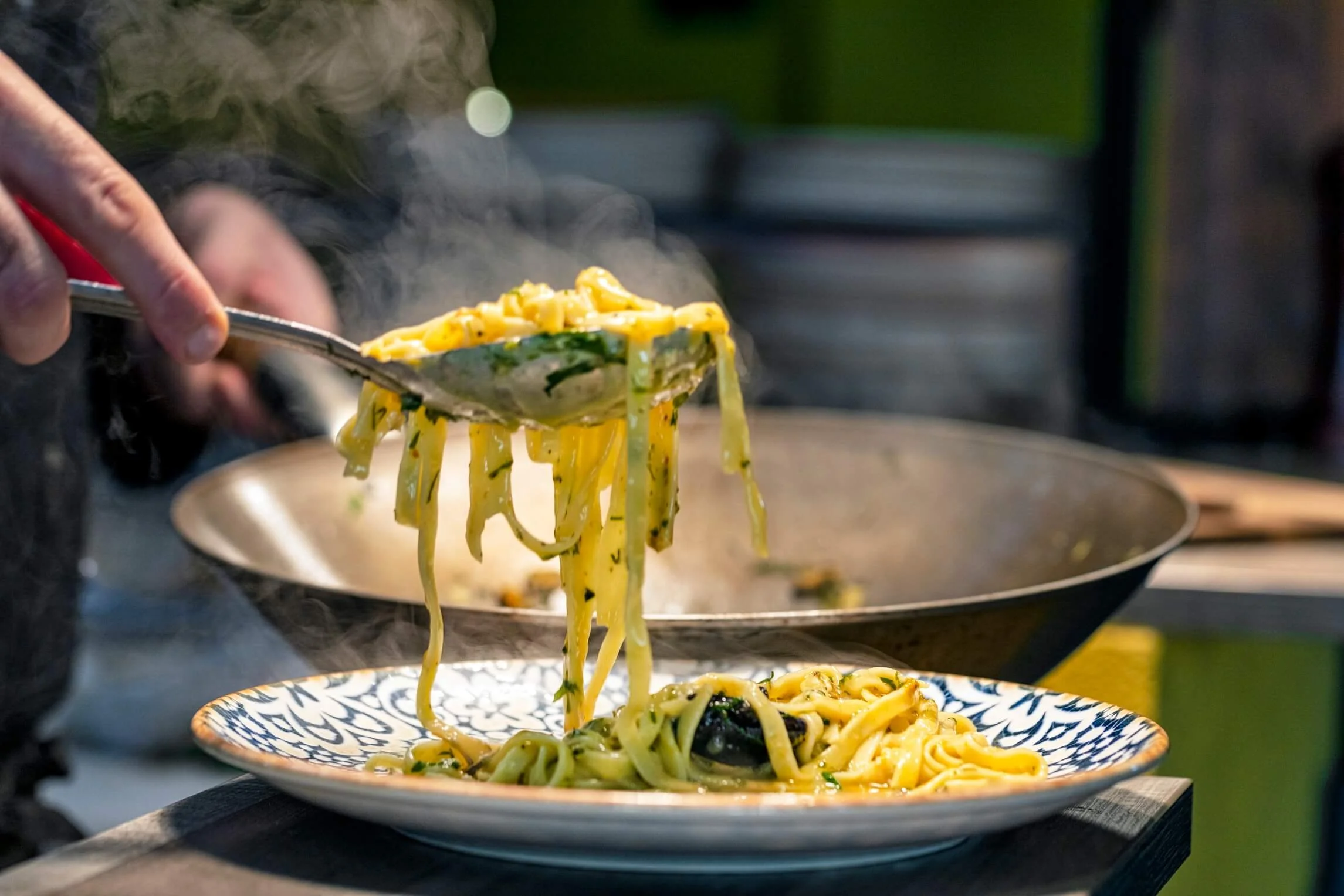 Close-up of a person serving steaming hot pasta with herbs onto a decorative plate.