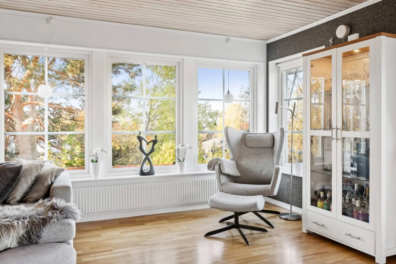A cozy living room corner with large windows showing a view of trees with fall foliage, a modern gray armchair, a white display cabinet filled with bottles, a tall white floor lamp, and decorative vases with white flowers.