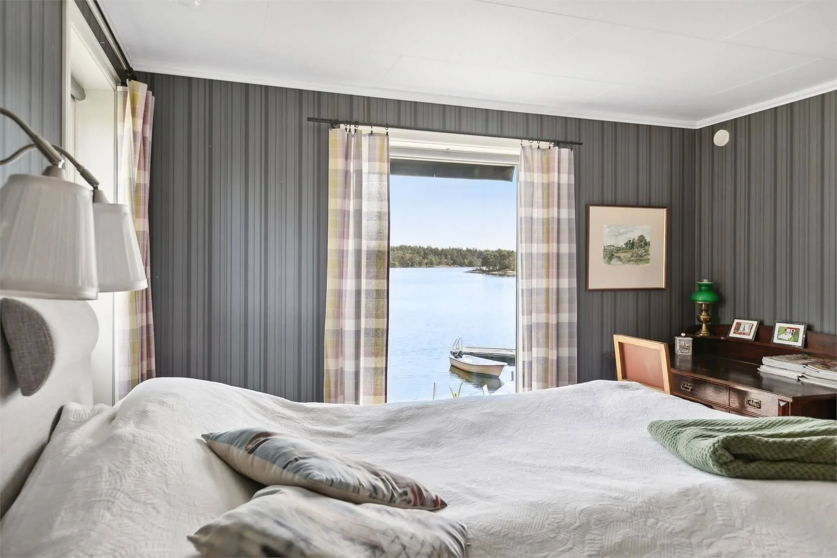 A bedroom with a large window showing a view of a boat on a lake and trees in the background. The room has striped gray walls, white bedding, and a wooden side table with framed pictures and a green lamp.