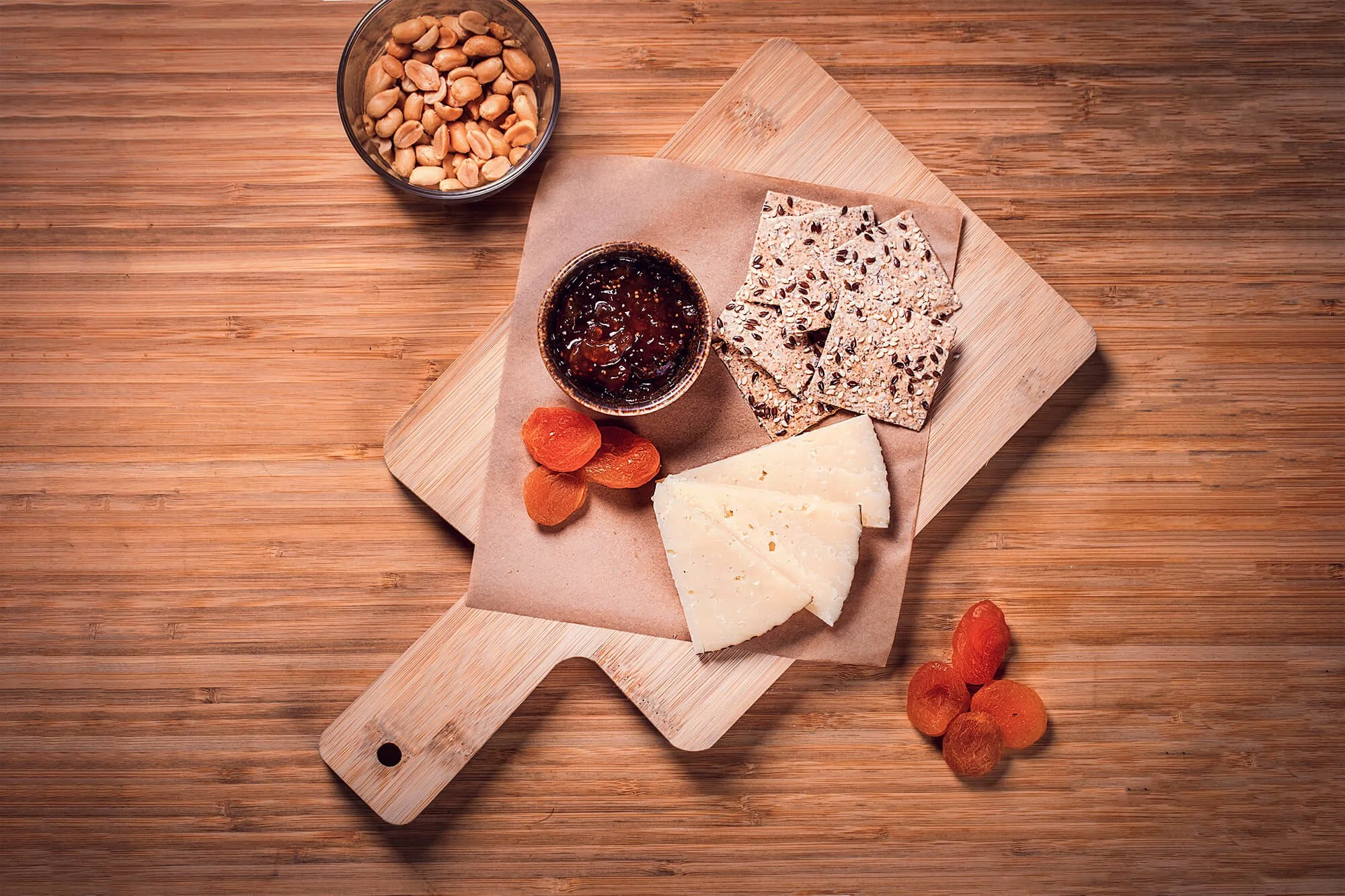 A cheese platter on a wooden surface. It includes three slices of white cheese, a small bowl of dark jam or jelly, dried apricots, a block of multigrain crispbread, and a black bowl filled with peanuts.