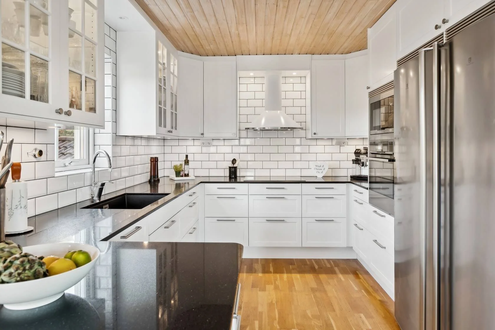 Modern white kitchen with black countertops, stainless steel appliances, white subway tile backsplash, wooden ceiling, and wooden floor.
