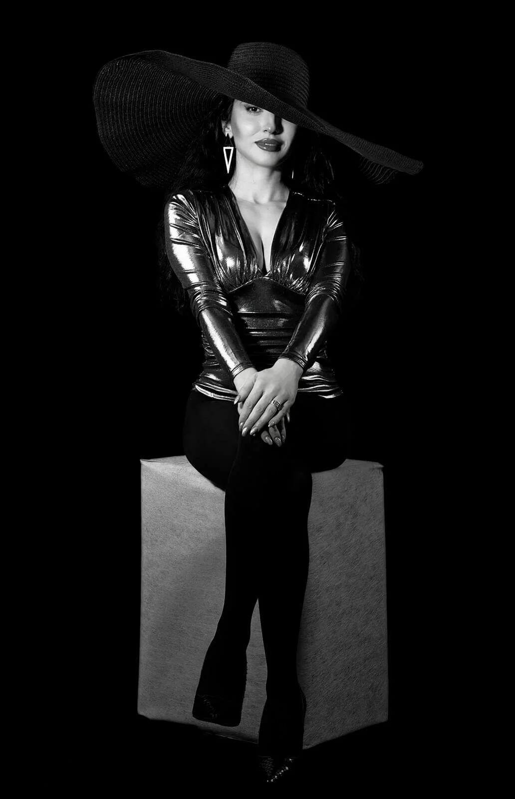 Black and white photo of a woman sitting on a cube, wearing a shiny black top, black pants, and large wide-brimmed hat, with earrings and lipstick, against a black background.