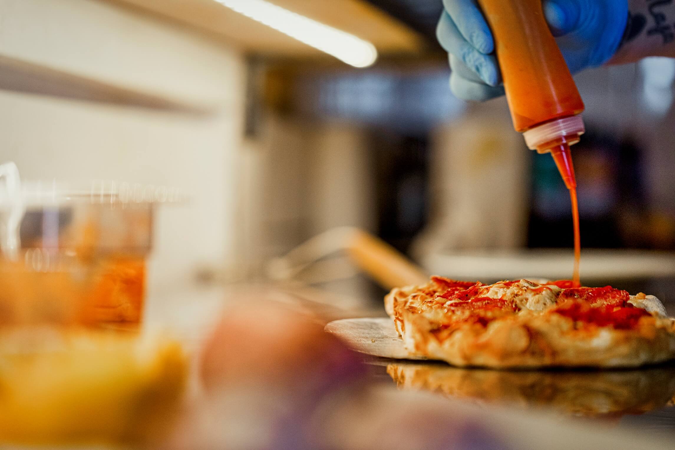 A person wearing a blue glove is squeezing orange sauce onto a freshly baked pizza. The pizza is on a wooden pizza peel, and the scene appears to be in a kitchen or pizzeria.