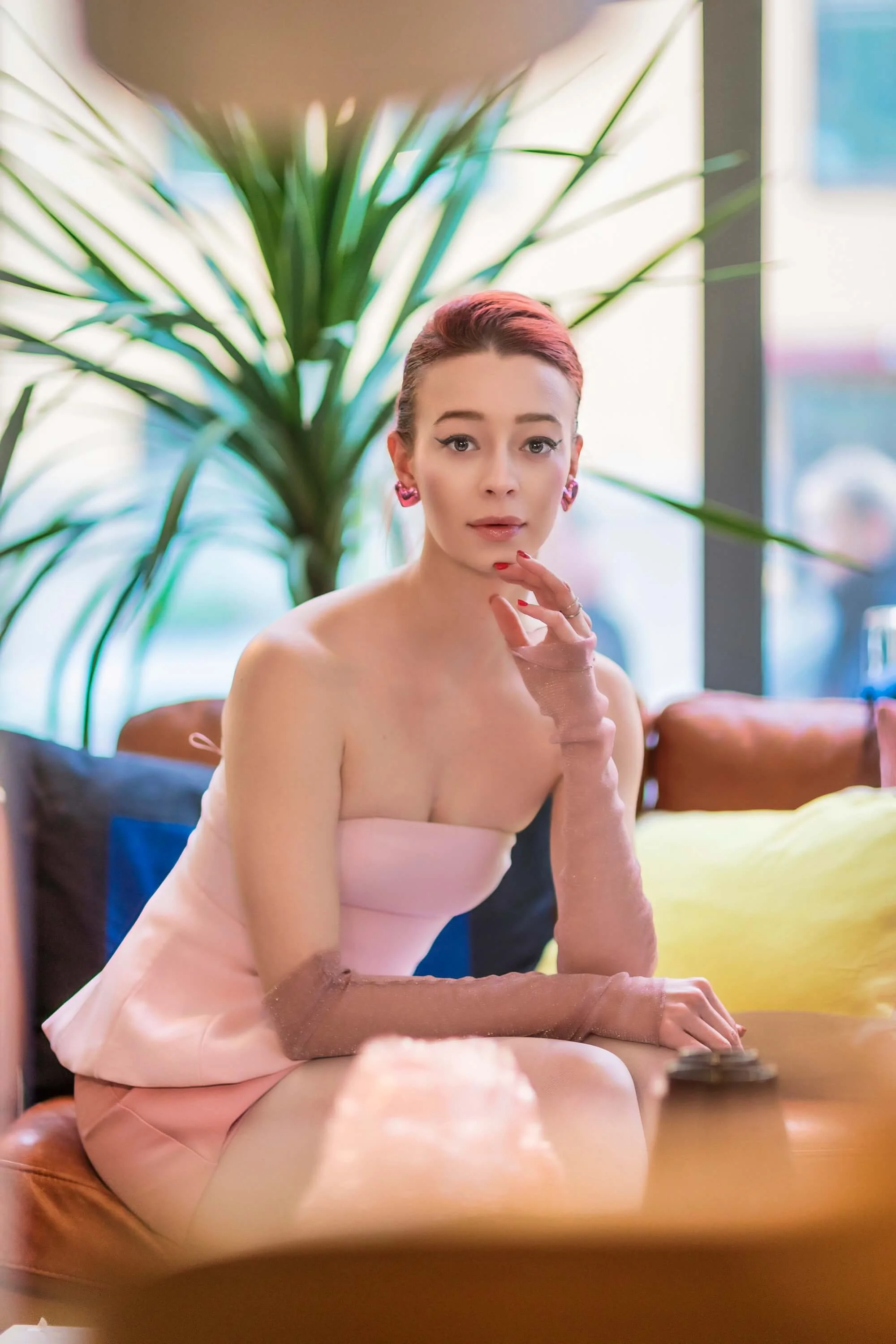 A young woman with short red hair and pink earrings sitting indoors near a large plant with green leaves, wearing a strapless pink dress with long matching gloves, and looking directly at the camera.