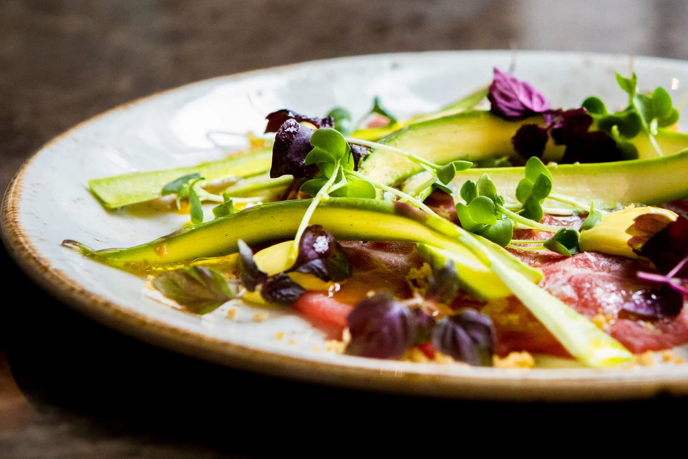 A close-up view of a plate with sliced avocado, microgreens, and edible purple flowers, garnished with a dressing or sauce.