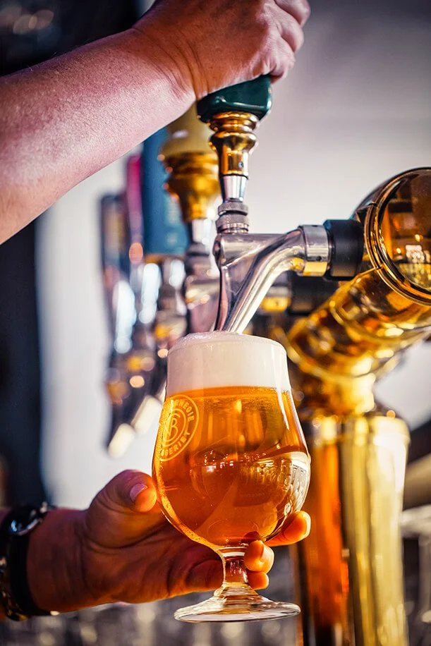 A person is pouring a glass of craft beer from a tap at a bar; the beer has a foamy head and a golden color.