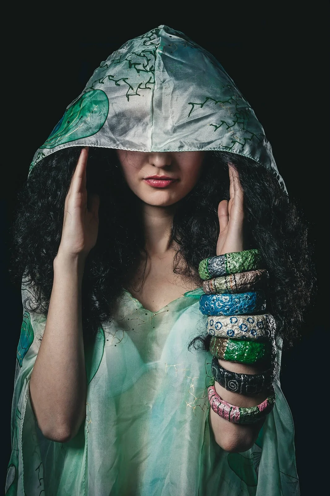 A woman with long curly black hair wearing a hooded garment with green, blue, and black abstract patterns, holding her head with one hand and displaying multiple colorful textured bracelets on her wrist against a dark background.
