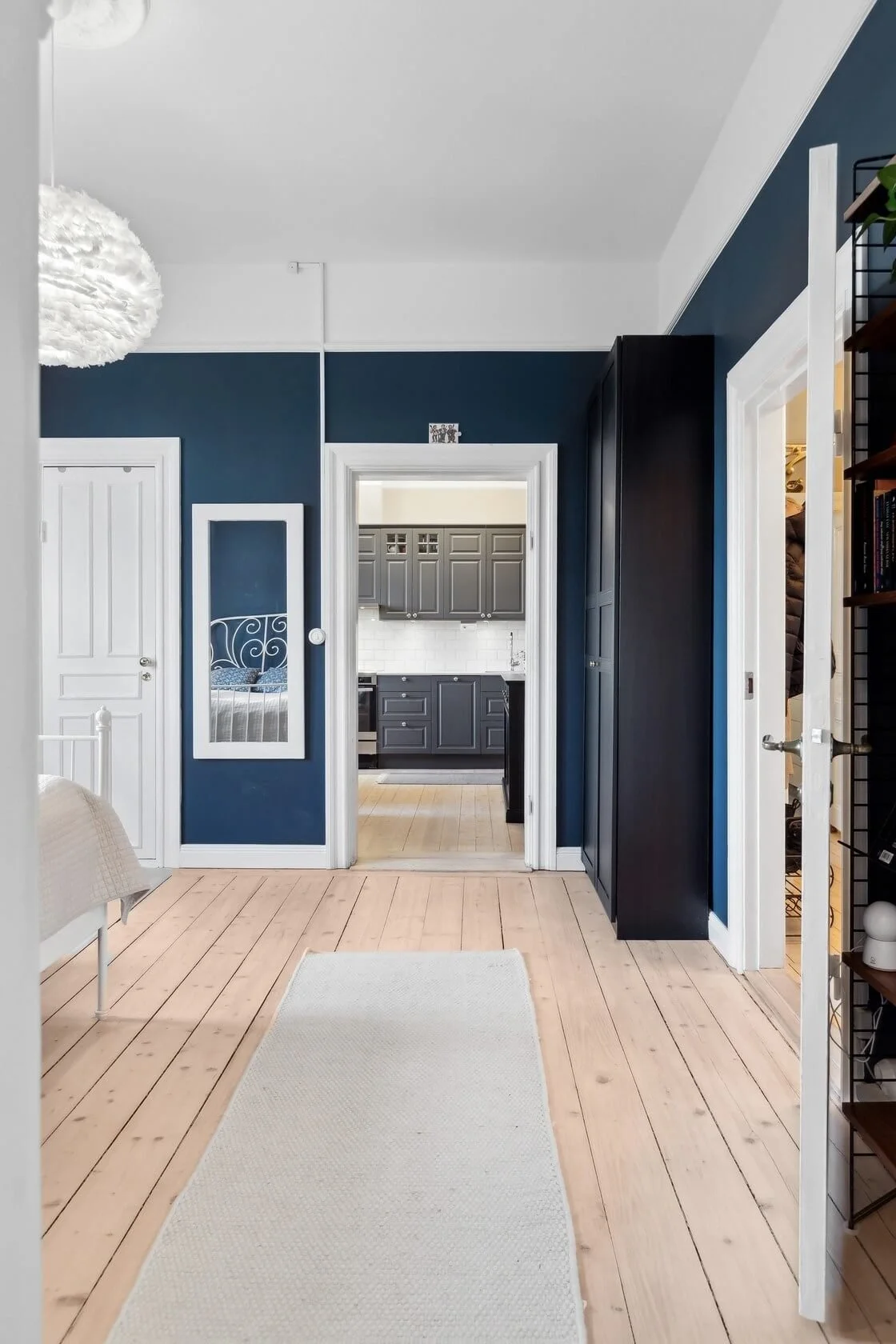 View of a room with light wood flooring, dark blue walls, and white trim. A doorway leads to a kitchen with gray cabinets. A white bed frame is partially visible on the left, and a white rug lies on the floor.
