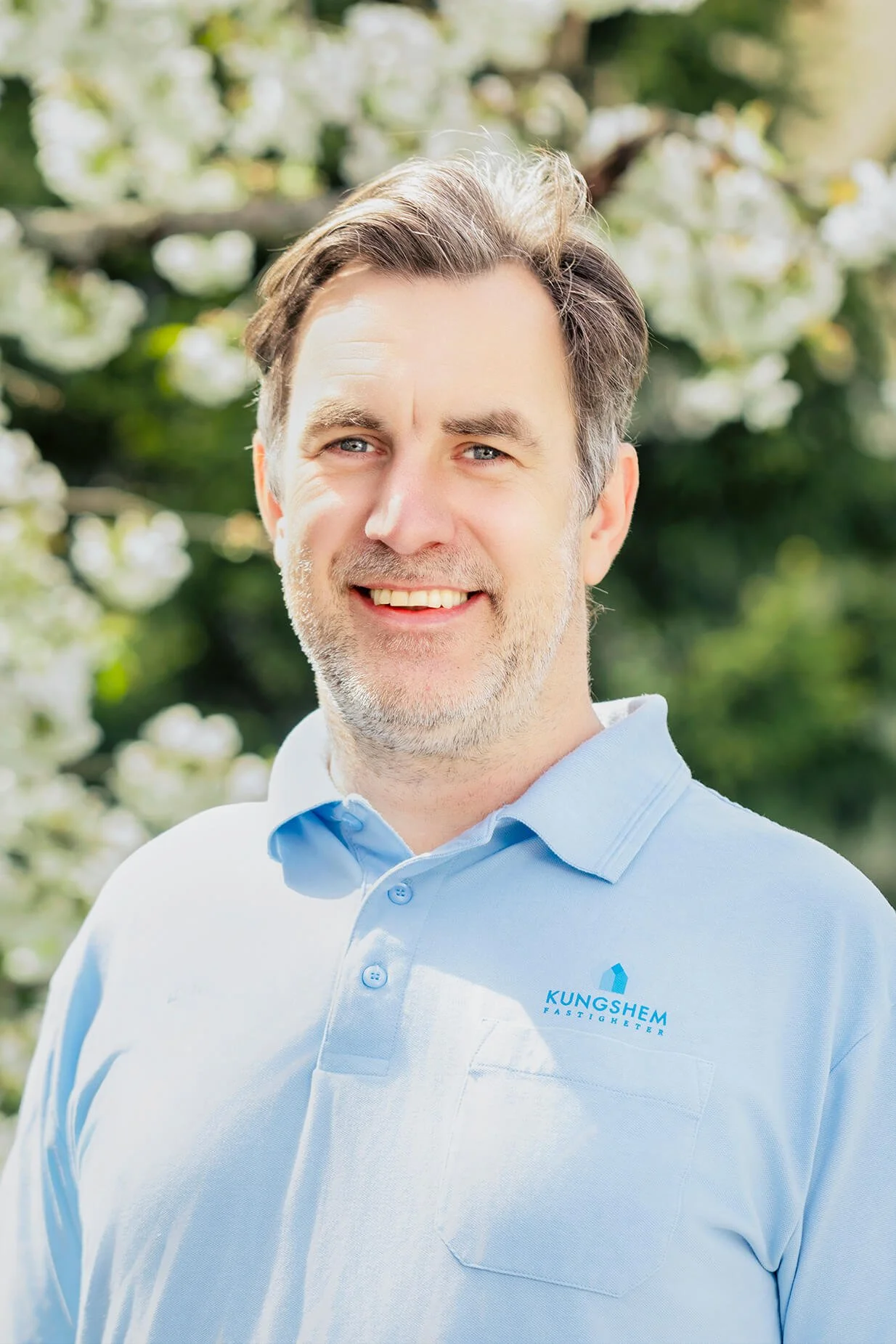 A smiling man with gray hair and a beard, wearing a light blue polo shirt with a "KungShem Fastigheter" logo, standing outdoors with blurred greenery and white blossoms in the background.