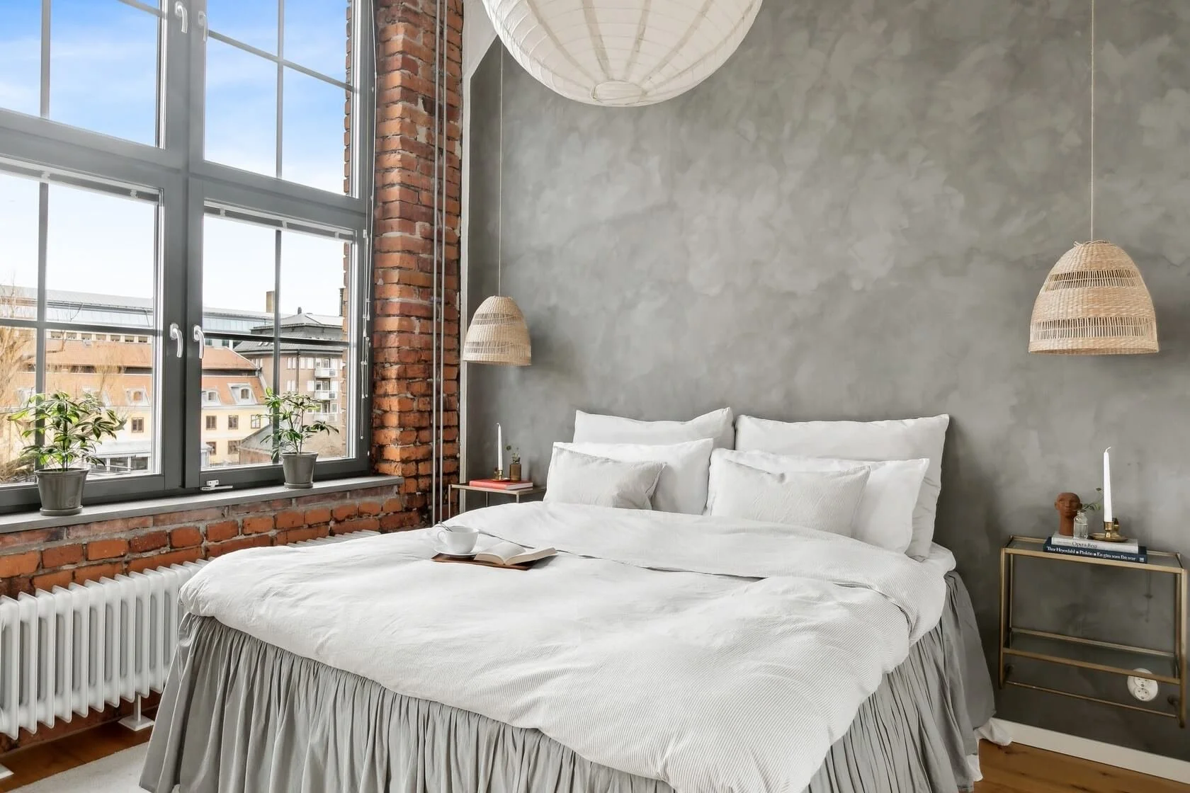 A cozy bedroom with a large window, exposed red brick wall, concrete wall, and a white bed with pillows. There are two hanging wicker lamps, and small side tables with decor items.